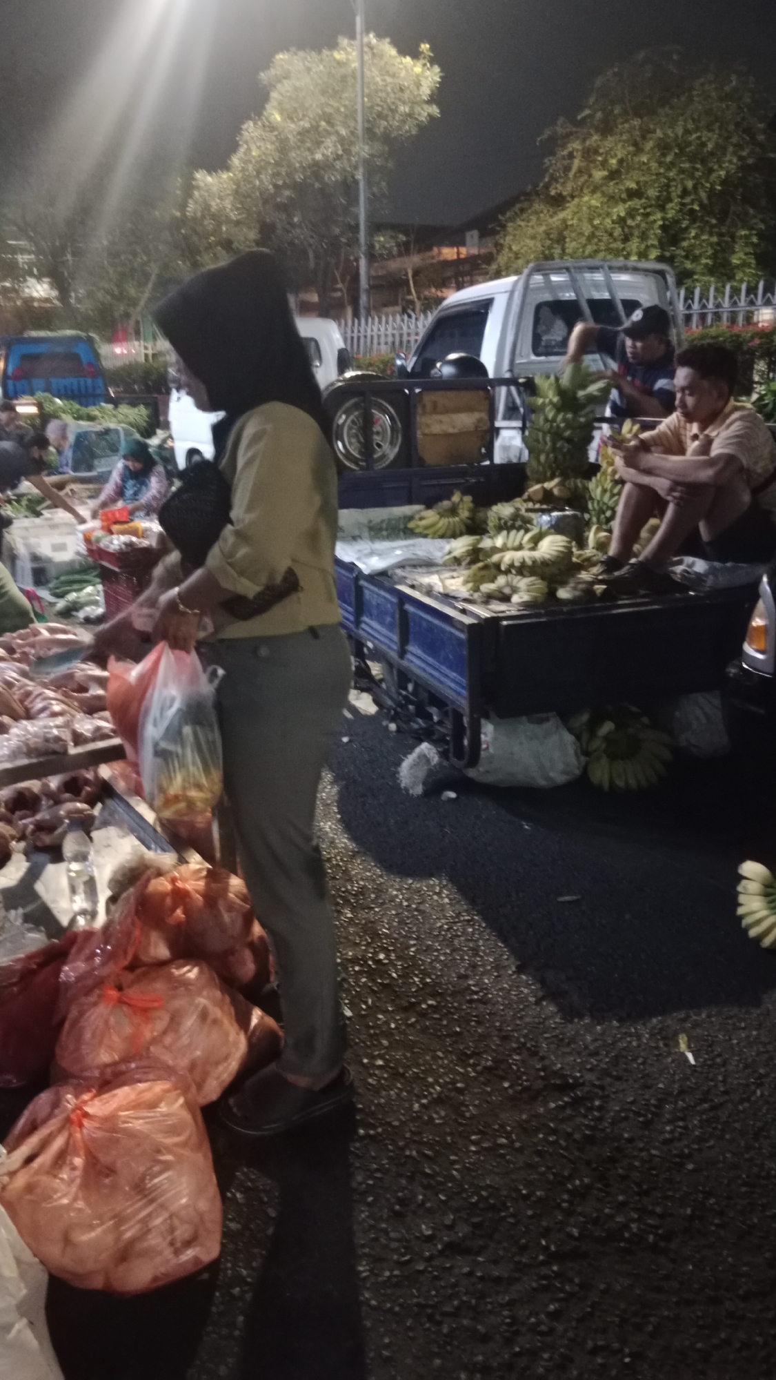 The image depicts a street market at night in Semarang, Indonesia. The foreground features a person wearing a hijab and a light-colored shirt, holding plastic bags filled with goods. Next to them are large, translucent plastic bags containing what appears to be produce or other items, tied with red string. A truck with a blue flatbed is visible behind the person, loaded with bunches of bananas. Two men are seated on the truck, one wearing a dark shirt and cap, and the other in a light-colored shirt. Other stalls and vehicles are present in the background, with streetlights illuminating the scene. The ground is asphalt, and the overall atmosphere suggests a bustling, nighttime market.