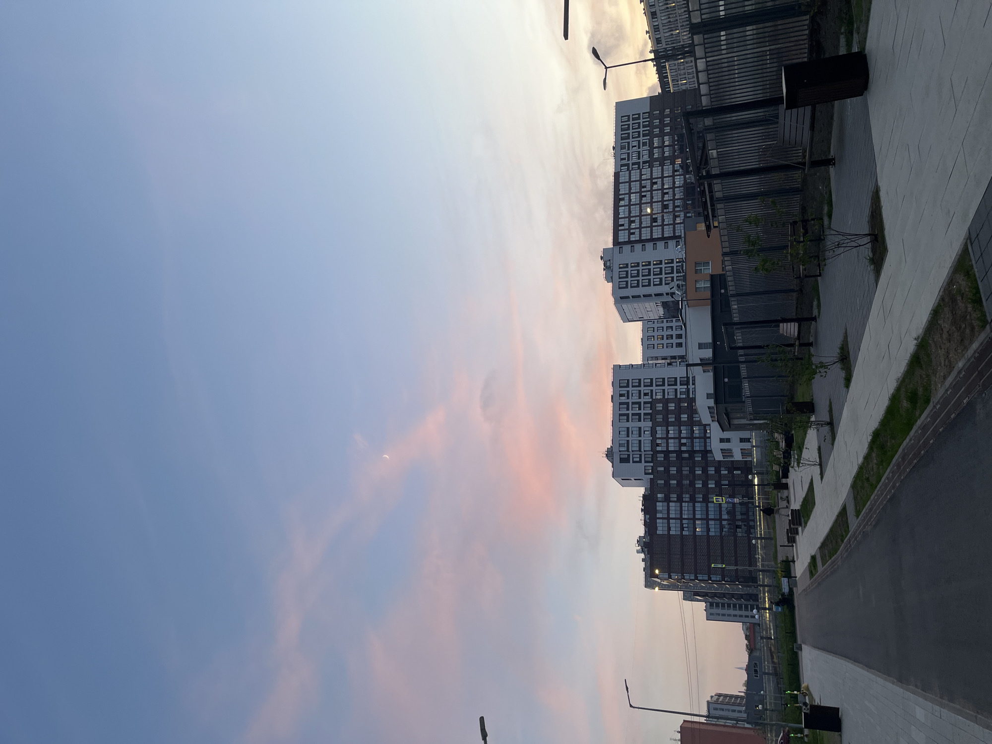 The image shows a street scene in what appears to be a newly developed urban area. Multiple modern, high-rise apartment buildings line the right side of the frame, constructed with dark brick and white accents. A paved path runs parallel to the buildings, with occasional benches and light posts along the way. The sky occupies the left side of the image, displaying soft pink and blue hues. There is a crescent moon in the upper left corner. The image appears to be rotated 90 degrees counter-clockwise. This is likely Saint Petersburg, Russia based on the architectural style and urban planning.