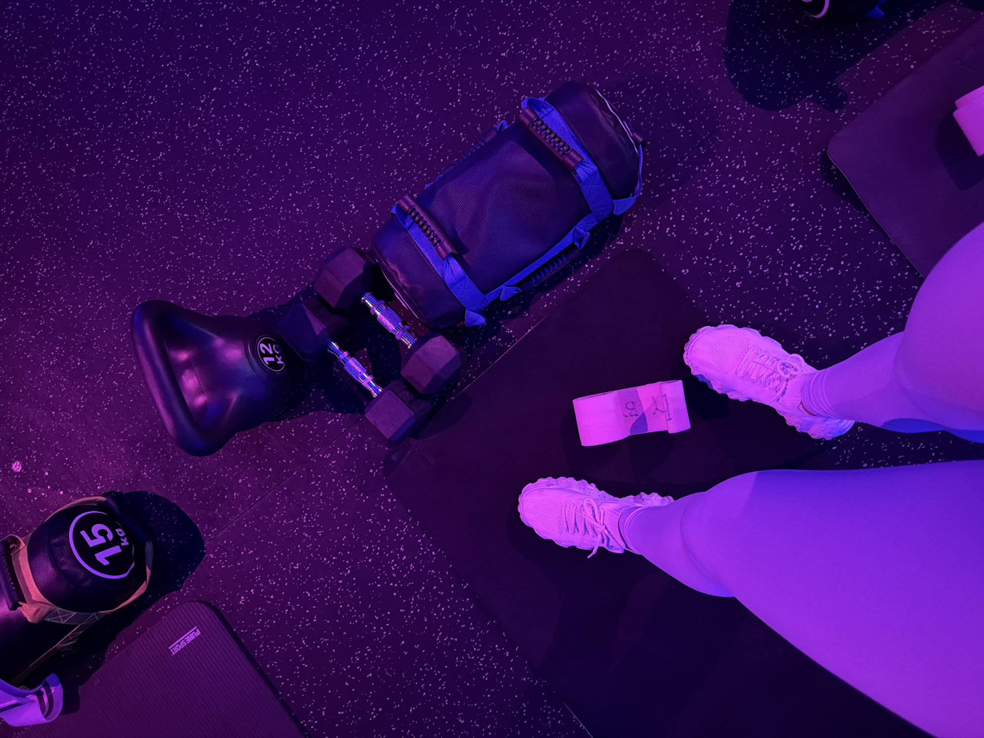 This is an eye-level, over-the-shoulder shot of a person's legs and feet wearing white sneakers and light purple leggings, standing on a black workout mat in what appears to be a gym. The gym floor is black with white speckles. To the right of the person, there is a black weightlifting bag with blue straps and handles, and two dumbbells. Further to the left, a black kettlebell with a 12kg weight indicator is visible, as well as another kettlebell with a 15kg weight indicator. A pink resistance band is on the mat near the person's feet. The lighting in the room is predominantly purple. This image was not taken in Ko Samui District, Thailand.