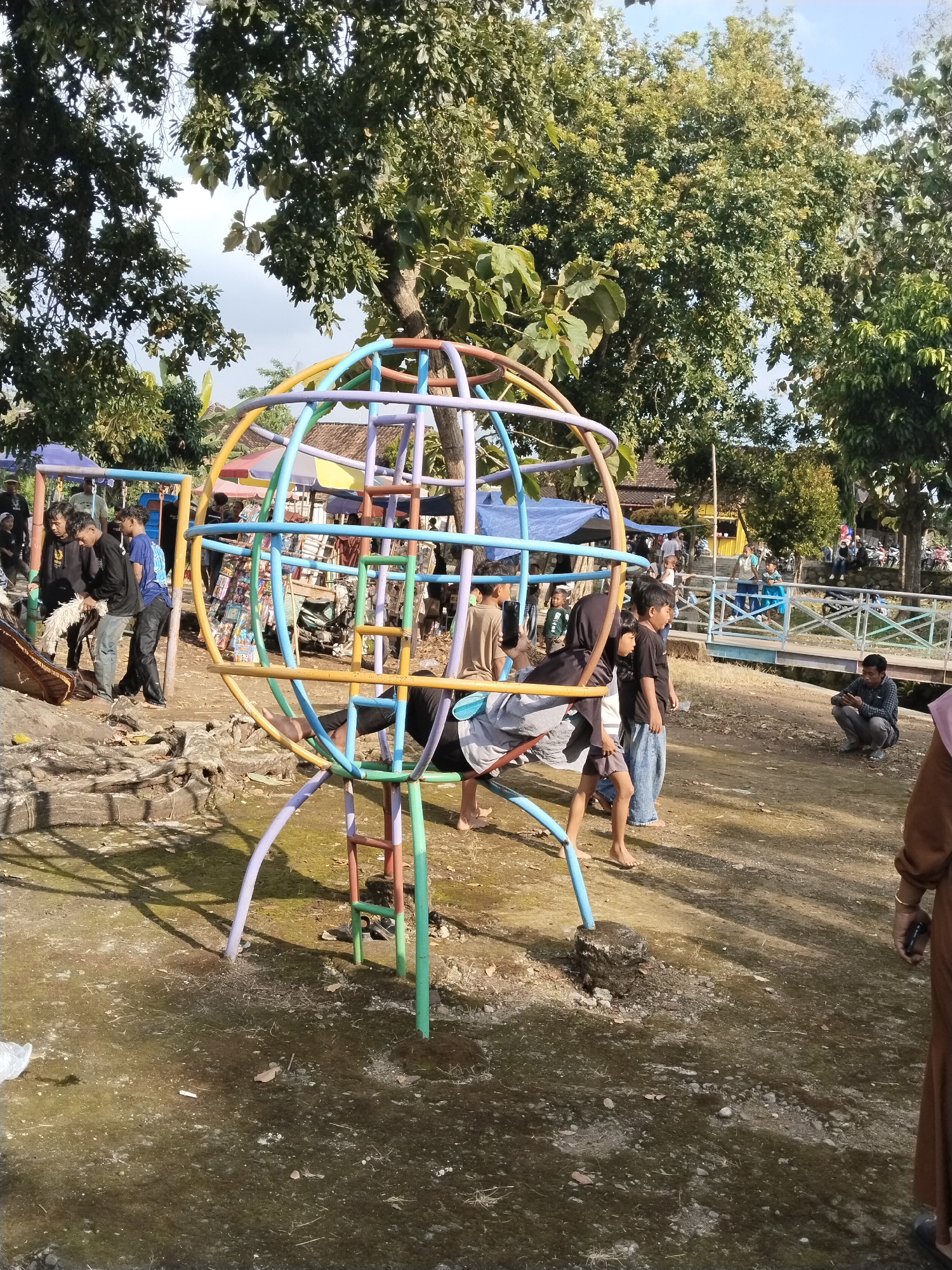 The image shows a public playground in Mojodoyong, Indonesia. A multi-colored, globe-shaped climbing frame dominates the scene. Several children interact with the structure. Some are climbing, while others stand nearby. In the background, other people are seen standing around a makeshift market area with stalls under blue tarps. A small bridge crosses a narrow waterway, and additional people are visible near the bridge. The ground is mostly bare earth with patches of moss and grass. Trees surround the playground, providing shade. The lighting suggests a sunny day. One man squats down and is on his phone to the right of the playground.