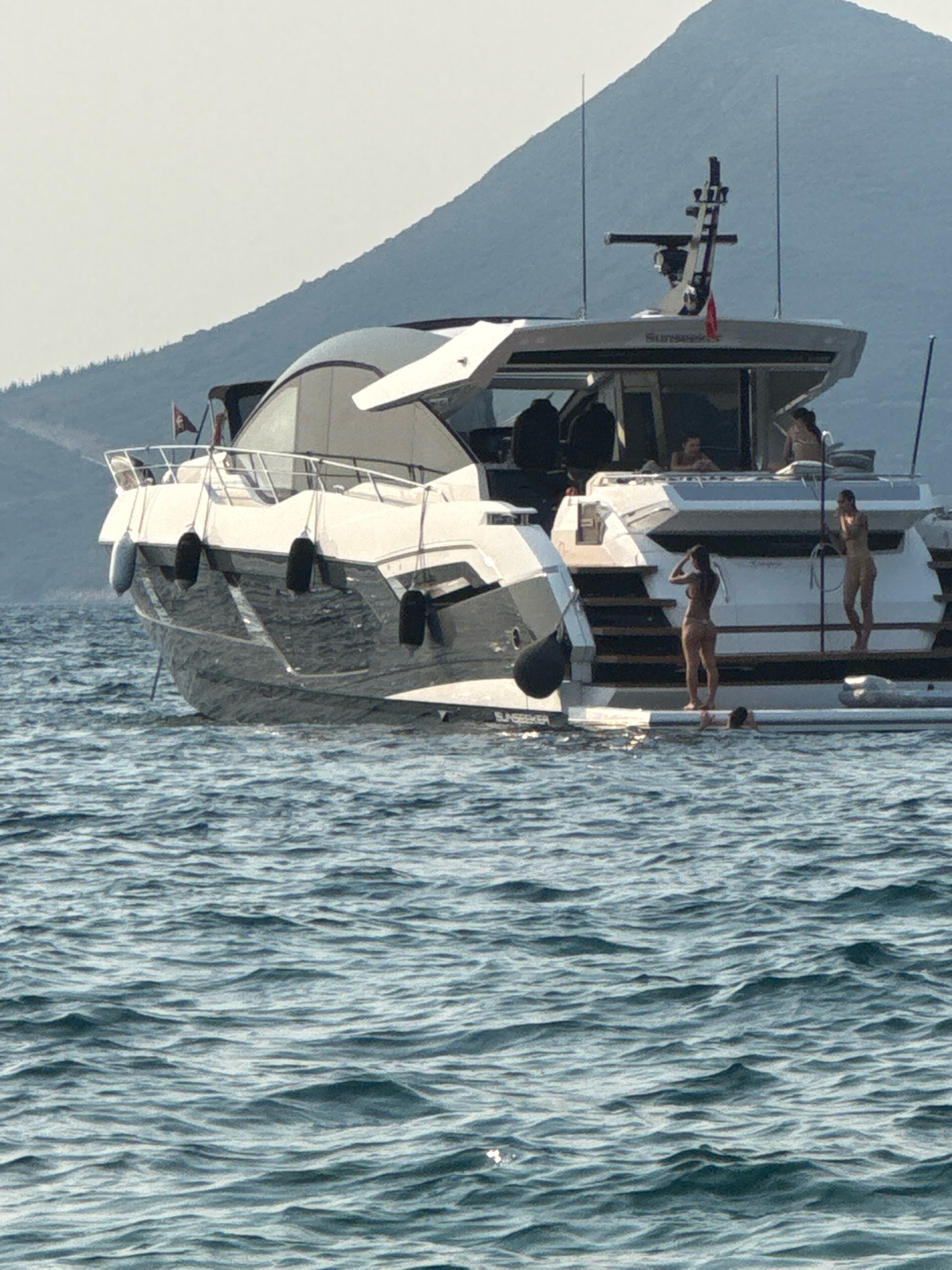 In the waters of Luštica, Montenegro, a large white yacht is anchored. The yacht has multiple levels and is equipped with various amenities for leisure. Several individuals are visible on the yacht, some appearing to be sunbathing or relaxing on the deck, while others are in the water. Two women in bikinis are standing on the aft deck of the yacht, one in a brown bikini and the other in a light-colored bikini, facing away from the camera. Another person, also in swimwear, is partially submerged in the water near the stern of the yacht. The background features a hazy, mountainous landscape typical of the Montenegrin coast. The water is choppy, indicating a moderate breeze. The scene conveys a sense of a leisurely day spent at sea.