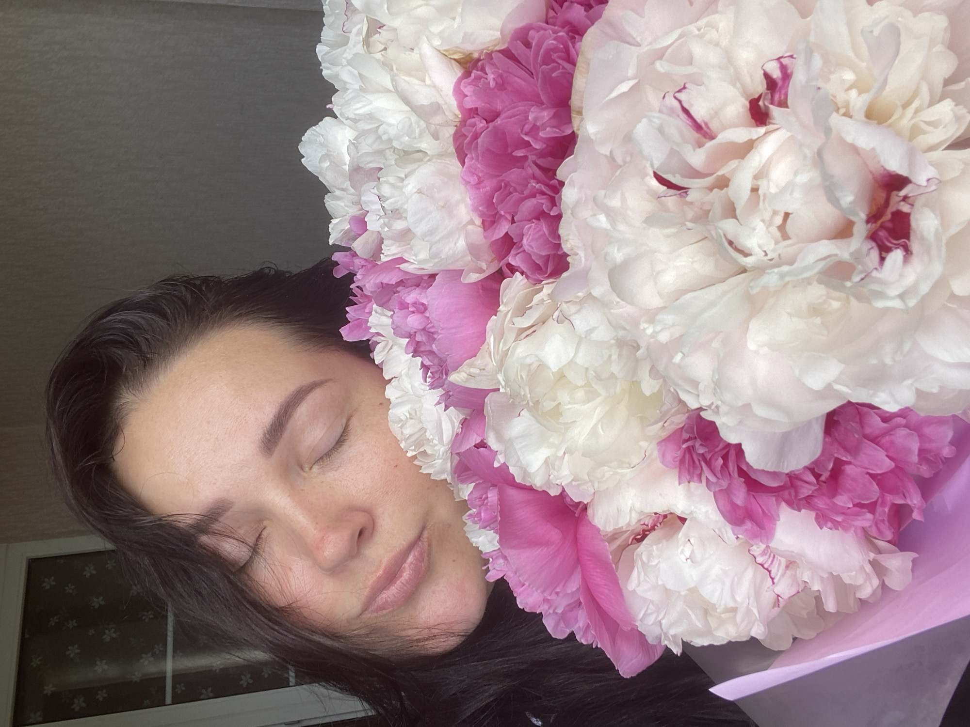 A woman with closed eyes is holding a large bouquet of white and pink peonies. The woman has dark hair and light skin. She is wearing a dark shirt. The bouquet is wrapped in light purple paper. The scene is lit by soft, natural light. A window is visible in the background. It is unclear whether the location is Sochi, Russia, without additional information.