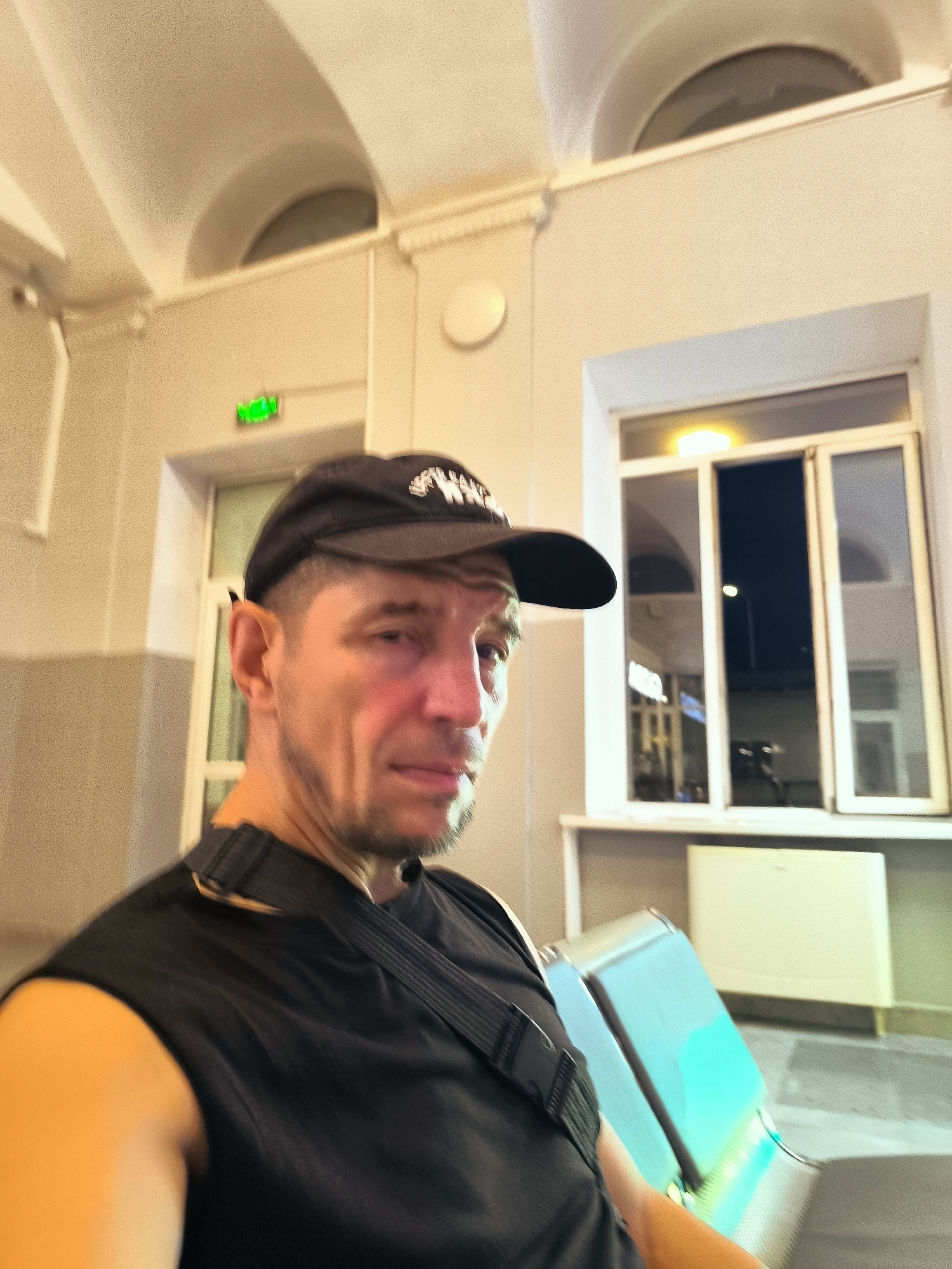 A man wearing a black baseball cap and a black sleeveless shirt is seated on a bench, looking to his right. He has short, light brown hair, a light beard, and appears to be in his late 30s or early 40s. He is wearing a black strap across his chest. The background shows a waiting area with architectural features like arched ceilings and a large window. A green exit sign is visible above a doorway in the distance. The bench he is sitting on has a metallic finish with a blue-green reflective surface. The overall lighting suggests it is either evening or indoors. The location is identified as the city administration of Qarağandı, Kazakhstan.