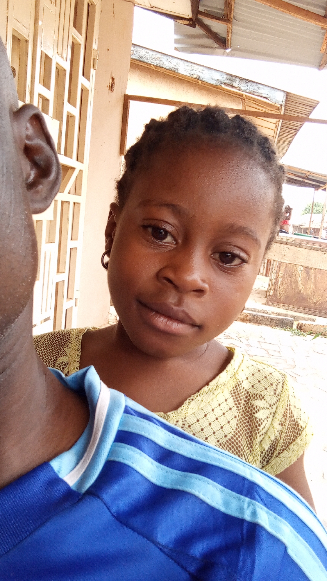 A girl with braided hair and dark skin is looking directly at the camera with a neutral expression. She is wearing a light yellow lace top. To her left is the side of a man's head and his shoulder, wearing a blue soccer jersey with lighter blue stripes and a white collar. The background is slightly out of focus but shows a building with a white patterned wall and a corrugated metal roof. The location is determined to be Sabon Tasha, Nigeria.