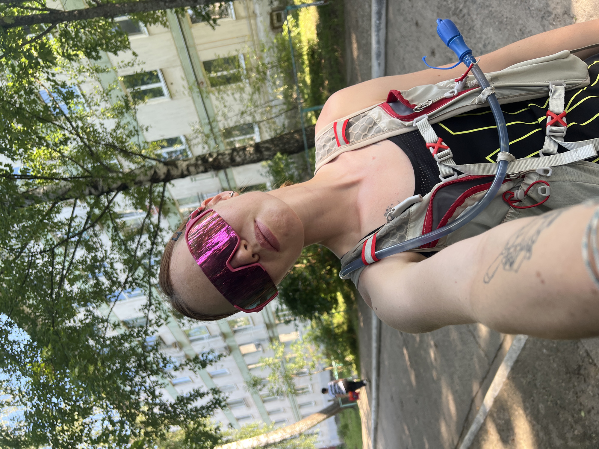 A person is taking a selfie outdoors on a sunny day in Chaikovsky, Russia. The person is wearing large, reflective pink sunglasses and a hydration vest. The background shows a light-colored, multi-story apartment building with green trim and numerous windows, characteristic of Soviet-era architecture. Trees with lush green foliage are present in the foreground and between the viewer and the building. A paved walkway or road is visible in front of the person, with a white line marking its edge. In the middle ground, to the left, another person is walking away from the camera, wearing a striped shirt and dark pants, and carrying a red object. The overall environment suggests a residential urban area with green spaces.