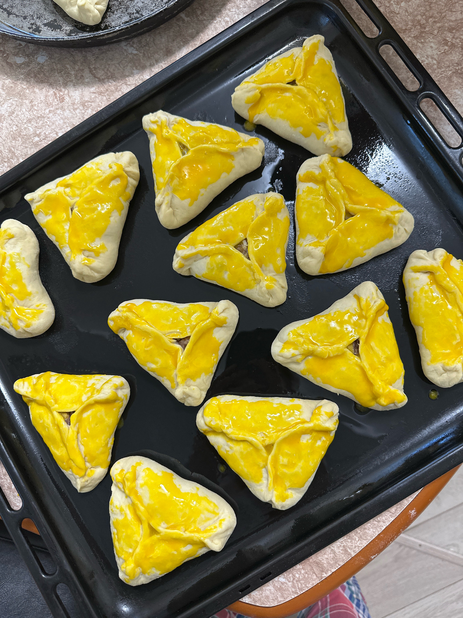 A black baking sheet is filled with approximately eleven triangular pastries topped with a yellow glaze. Some of the pastries are placed close together, while others have a small gap between them. The pastries are arranged in three rows on the baking sheet. The sheet itself appears to be slightly greasy and there are some droplets of liquid on its surface. The baking sheet is positioned on a speckled brown surface, and a portion of a red and blue plaid fabric is visible in the bottom right corner. This scene is likely occurring in a home kitchen in the city of Buinsk, Russia, given the context of its location.