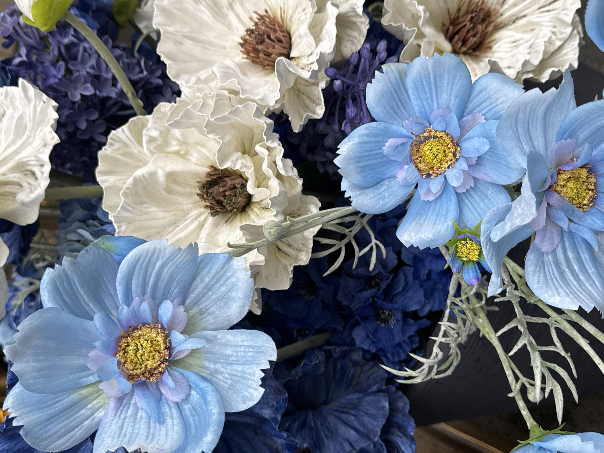 The image shows a close-up of an artificial flower arrangement. The flowers are a mix of white and blue, with some dark blue filler flowers. The white flowers have a brown center, and the blue flowers have a yellow center. The arrangement is dense and full. The background is dark and indistinct, which makes it difficult to determine the location. The arrangement appears to be resting on a dark surface. The image contains no people, actions or interactions. There is no evidence to suggest the location is Moscow, Russia.