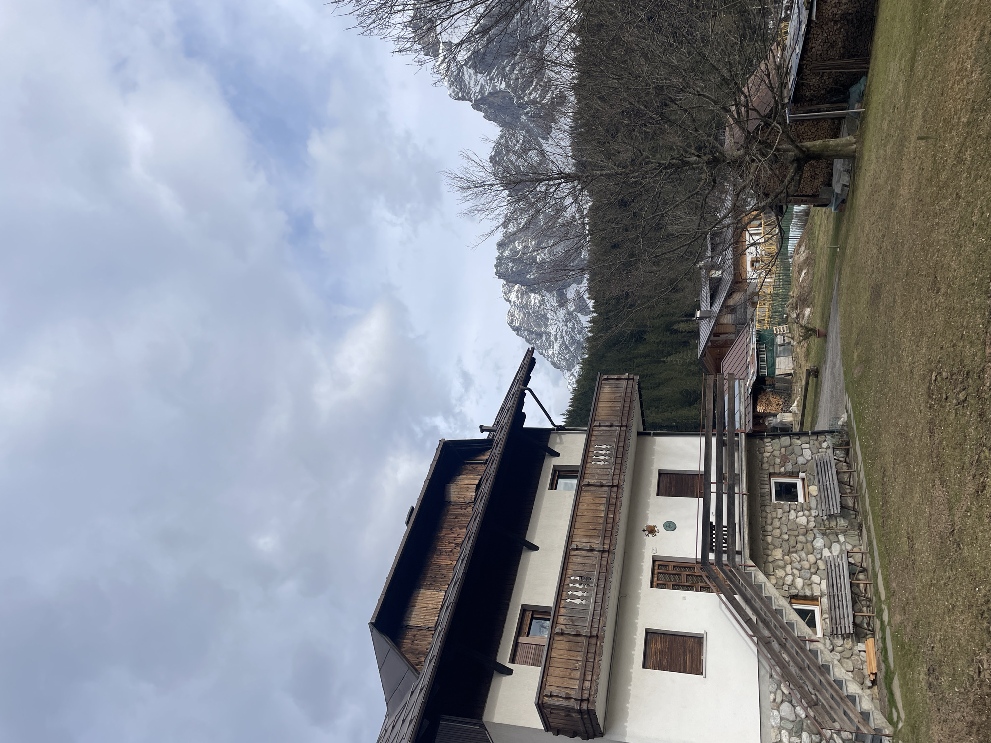The image shows a building in Sappada, Italy, with a mountain in the background. The building is multi-storied, with a wooden balcony on the upper floor. The lower part of the building has a stone facade. A set of stairs leads up to the entrance. The sky is cloudy.