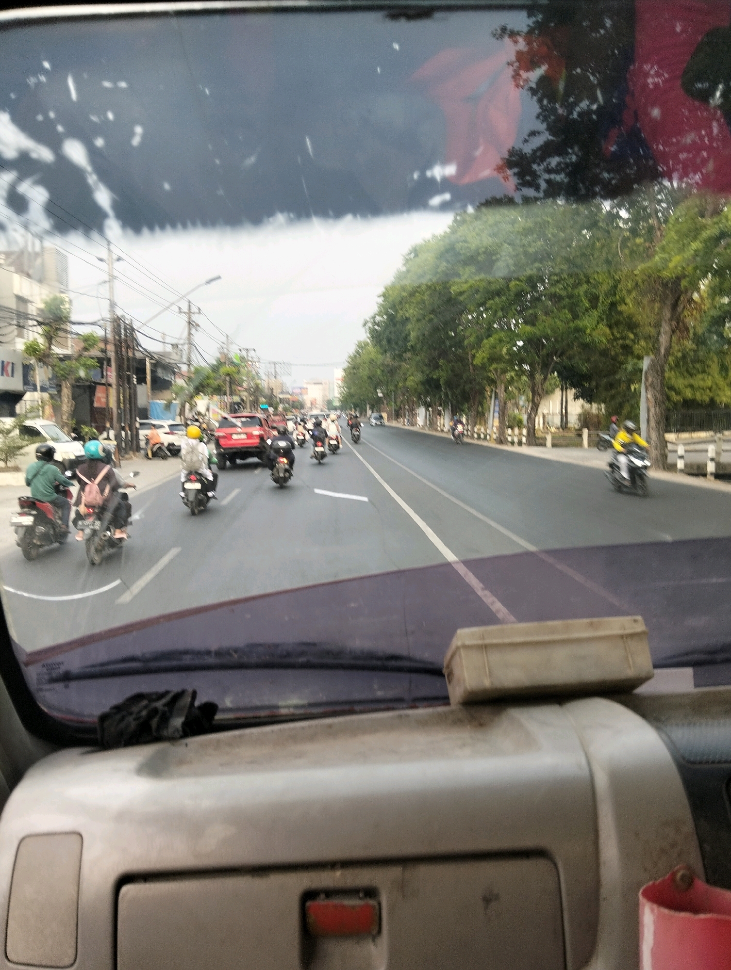 The image shows a view from inside a vehicle looking out through the windshield at a busy street scene in Semarang, Indonesia. The street is lined with trees and buildings, and there is a significant amount of traffic, primarily motorcycles. Several motorcycles are visible, carrying drivers and passengers, some wearing helmets. There are also cars and possibly other vehicles on the road. The sky appears overcast. The foreground shows the interior of the vehicle, including the dashboard and a piece of black cloth or fabric resting on it. A light-colored box is placed on top of the dashboard. The scene suggests a typical day in a bustling urban environment.