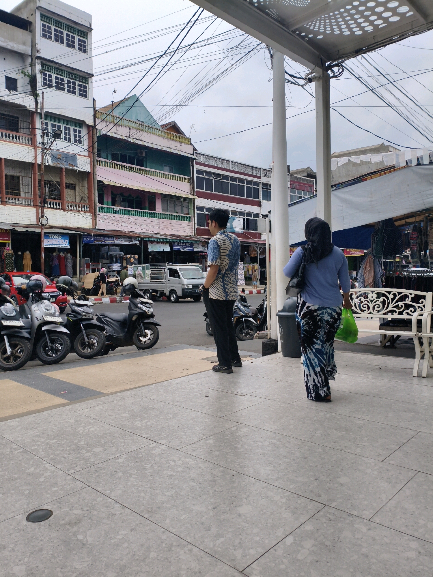 In the city of Unknown, Unknown, a street scene unfolds under an overcast sky. The foreground features a wide pedestrian walkway paved with large, light gray speckled tiles. To the left, a row of parked motorcycles, primarily black and silver, are lined up against the curb. Beyond the motorcycles, a red car is parked, and storefronts line the street. One storefront displays clothing and signage for "TOKO". The buildings are multi-story structures with a mix of architectural styles, some appearing older with white facades and decorative elements, while others have more modern, colorful exteriors in shades of pink and green. Overhead, a complex network of electrical wires crisscrosses the sky, suspended from poles.
Further into the scene, a man stands on the sidewalk, facing away from the camera, wearing a patterned blue and white shirt and dark pants. To his right, a woman walks away from the camera, wearing a black hijab, a light blue long-sleeved top, and a long, patterned skirt in shades of blue, black, and white. She carries a green plastic bag in her right hand. Behind her is a white wrought-iron bench. A dark gray trash receptacle is positioned next to the bench. A small silver truck is visible on the street, along with other parked vehicles and a few pedestrians in the background near the shops. A white awning with circular perforations covers part of the pedestrian area, supported by white columns.