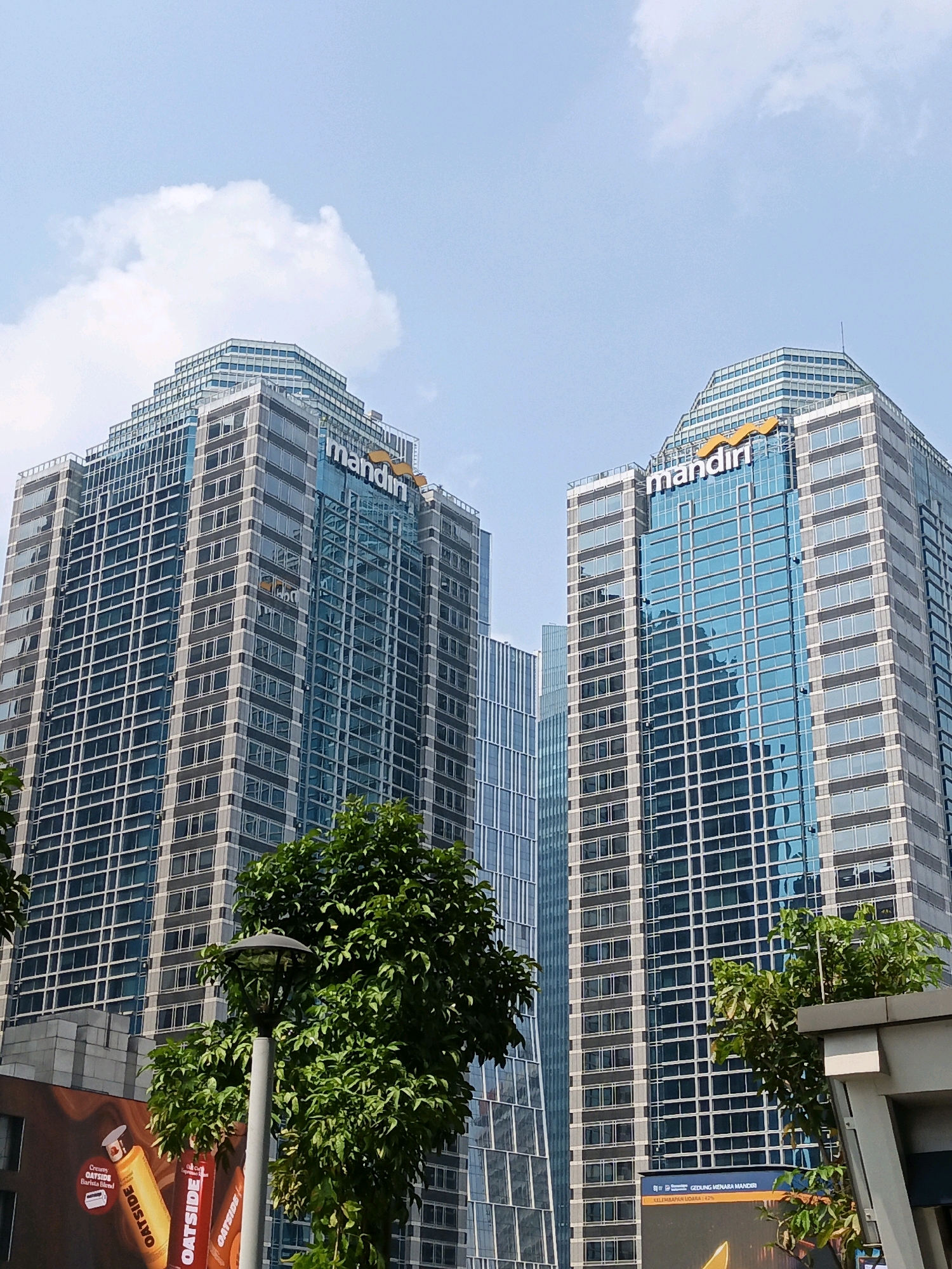 The image shows a low-angle, full-frame shot of two modern skyscrapers in Central Jakarta, Indonesia. Both buildings are tall and rectangular, with glass facades and visible grids of windows. The building on the left has a gray exterior with horizontal window panes, while the building on the right features blue-tinted glass. Both buildings display the "mandiri" logo in orange at their upper sections. In the foreground, lush green trees and a lamppost partially obscure the lower portions of the buildings. The sky above is a clear, bright blue with a few white clouds. The scene suggests an urban environment, likely a business district.