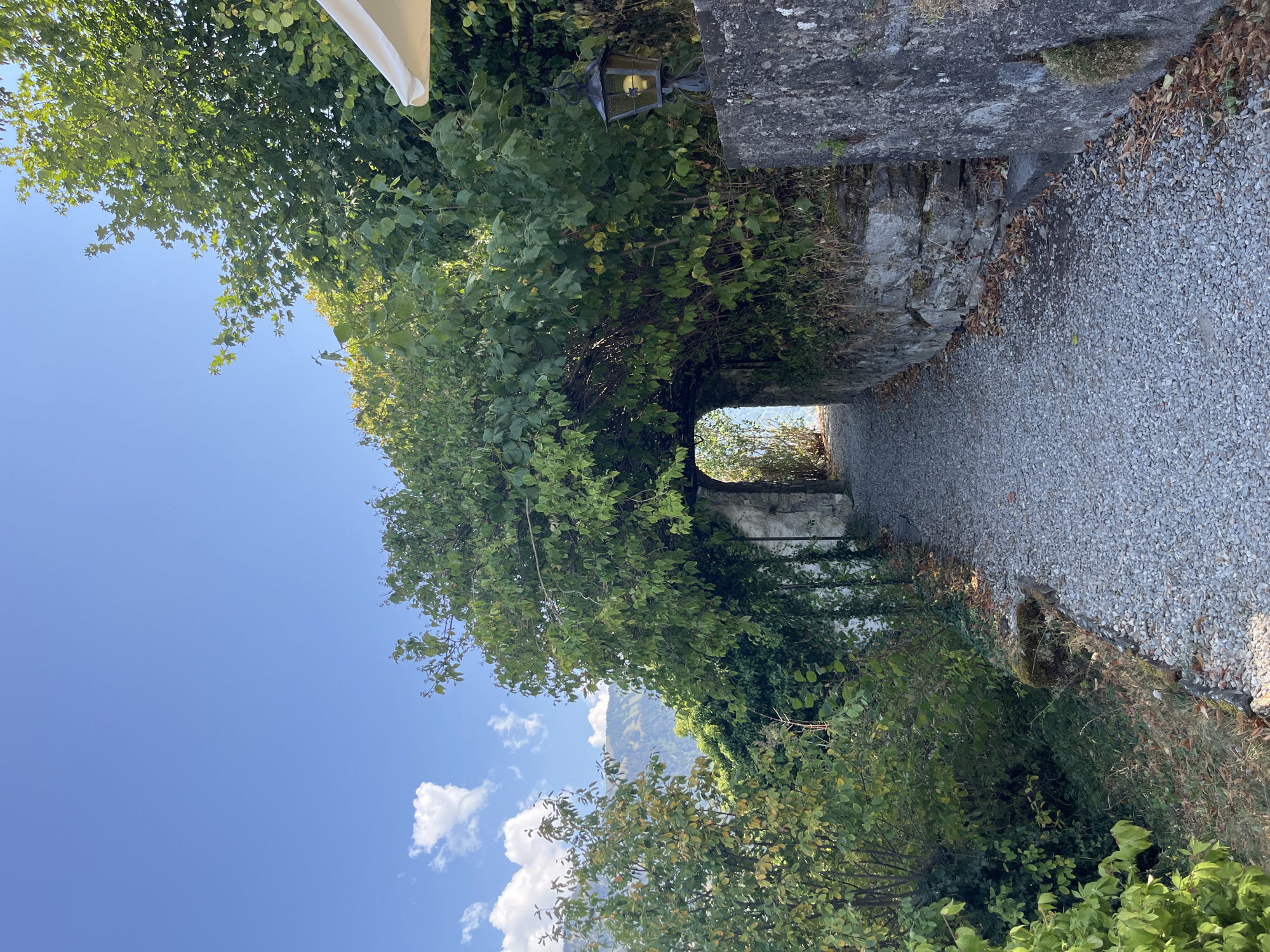 A sunlit, gravel pathway curves through a lush, green archway formed by overgrown trees and vines. The archway leads to a stone opening, beyond which a distant view of mountains under a clear blue sky with scattered white clouds is visible. To the right of the pathway, a stone wall is topped by a vintage-style lantern. To the left, dense foliage and trees border the path. The scene is set outdoors in a natural environment. The image is a vertical, full-shot composition. No people are present in the image. The location is identified as Epagny, Switzerland.