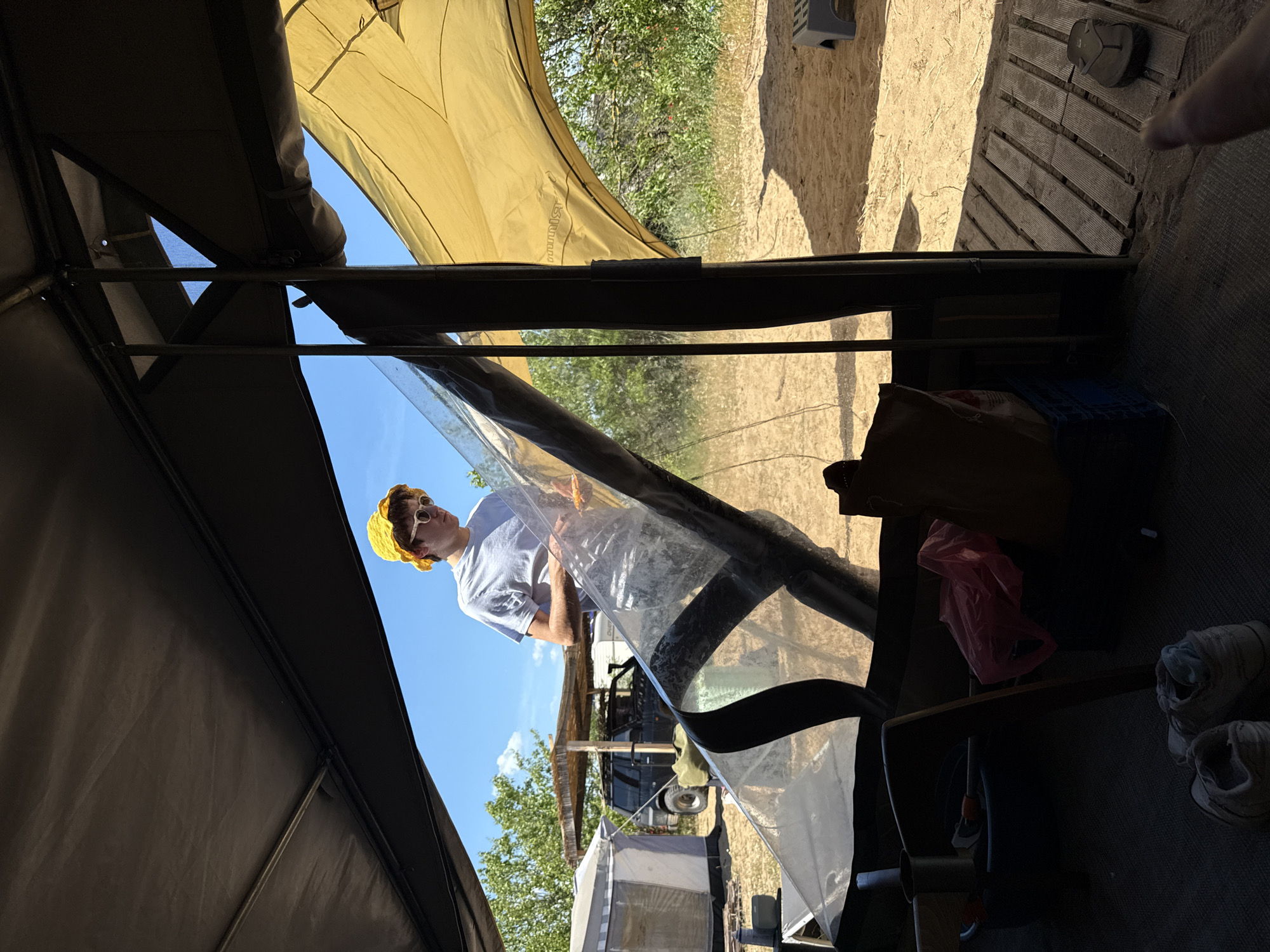 A low-angle shot depicts a partial view of a campsite interior, with an individual installing a clear vinyl awning. The person, wearing a yellow crochet hat and white sunglasses, is seen partially through the clear vinyl sheet. Outside the tent, there's a vehicle with gear on top and an awning, and another tent in the background. The tent's interior shows a variety of items: what looks like a blue milk crate, a pink grocery bag, a cardboard box, and a pair of sneakers. The ground is covered with what appears to be a wooden slat patio. The location appears to be a campsite near Sozopol, Bulgaria, but that is an estimation based on the environment and surrounding vegetation.