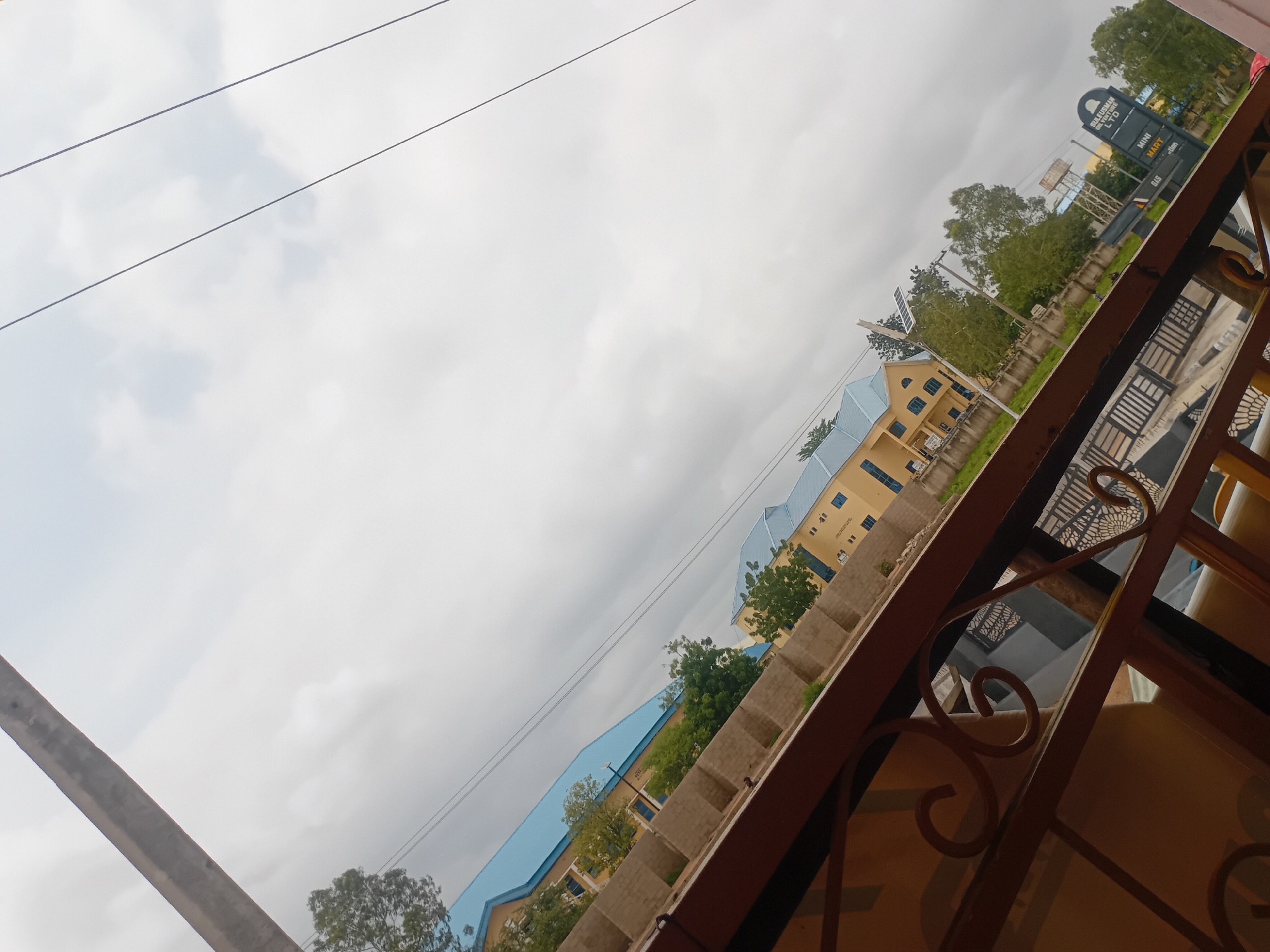 An eye-level, slightly tilted medium shot shows a street scene with a focus on buildings and a guardrail. The sky is overcast with a thick layer of gray clouds. In the foreground, a brown, ornate metal guardrail runs diagonally across the lower portion of the frame. Beyond the guardrail, a light brown brick wall is visible, and behind that, a yellow building with a blue roof. Further into the background, another yellow building with a blue roof is visible, along with several green trees. Power lines are visible in the upper portion of the image. On the right side of the frame, a sign for "SULEIMAN OIL AND GAS LTD MINI MART" is visible. The overall scene appears to be in a residential or commercial area.