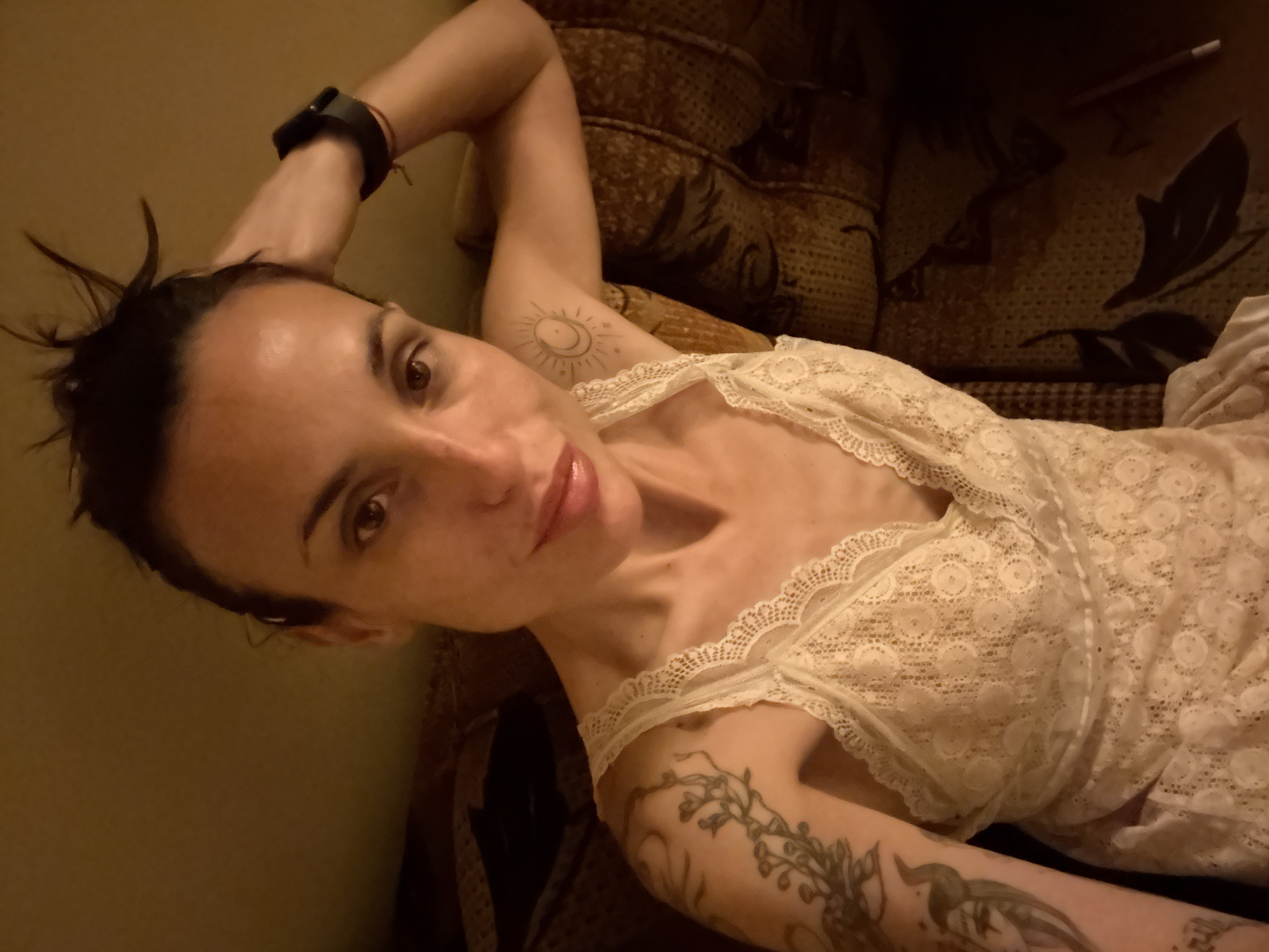 The image shows a woman in a light-colored, lace-trimmed camisole, seated on a patterned sofa. She has dark hair pulled up and is resting her right hand on her head, wearing a dark smartwatch on her left wrist. She has visible tattoos on her left arm, including a branch with leaves and a sun and moon design on her right arm. The setting appears to be indoors, with warm lighting. There is no information in the image to suggest a specific location in Pleven, Bulgaria.
