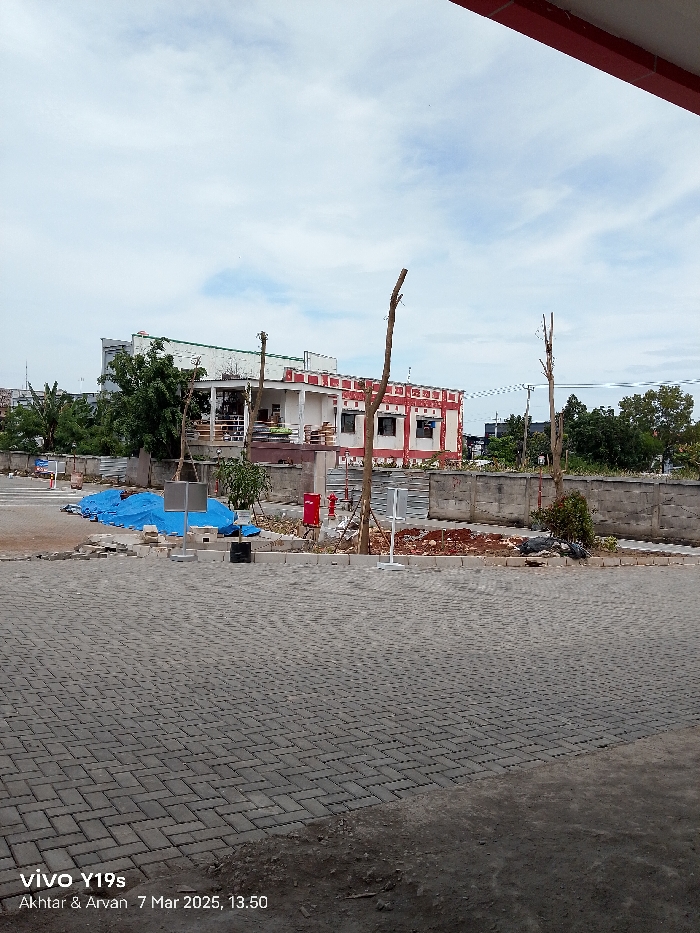 The image was taken in Indramayu, Indonesia at coordinates -6.343 latitude and 108.336 longitude. The scene shows a paved parking lot in front of a building under construction. There are several trees and shrubs around the parking lot, with a wall separating the parking lot from the building. There is a red fire hydrant near the wall and a blue tarp on the ground. The sky is clear with some clouds. The image was taken by a smartphone with a timestamp of March 7, 2025, at 1:50 PM. There are no people in the image.