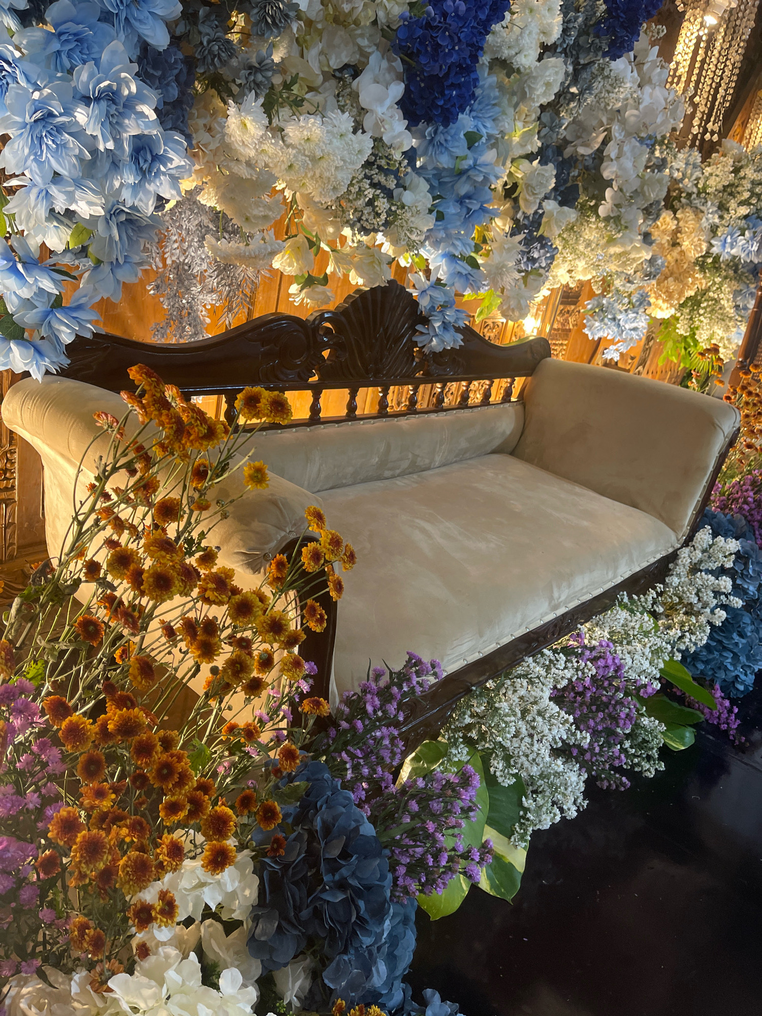The image depicts an ornate, velvet-upholstered chaise lounge positioned in front of a lush floral arrangement. The chaise lounge is a rich cream color with a dark, intricately carved wooden frame. The floral display is abundant, featuring a mix of blue, white, and yellow flowers cascading from above and surrounding the base of the lounge. Prominent amongst the lower blooms are clusters of vibrant orange and yellow chrysanthemums. The setting appears to be indoors, with wooden paneling visible behind the floral backdrop and a dark, reflective surface in the foreground, possibly a floor or table. The lighting is warm and somewhat dim, highlighting the textures of the flowers and the lounge. There are no people visible in the image.