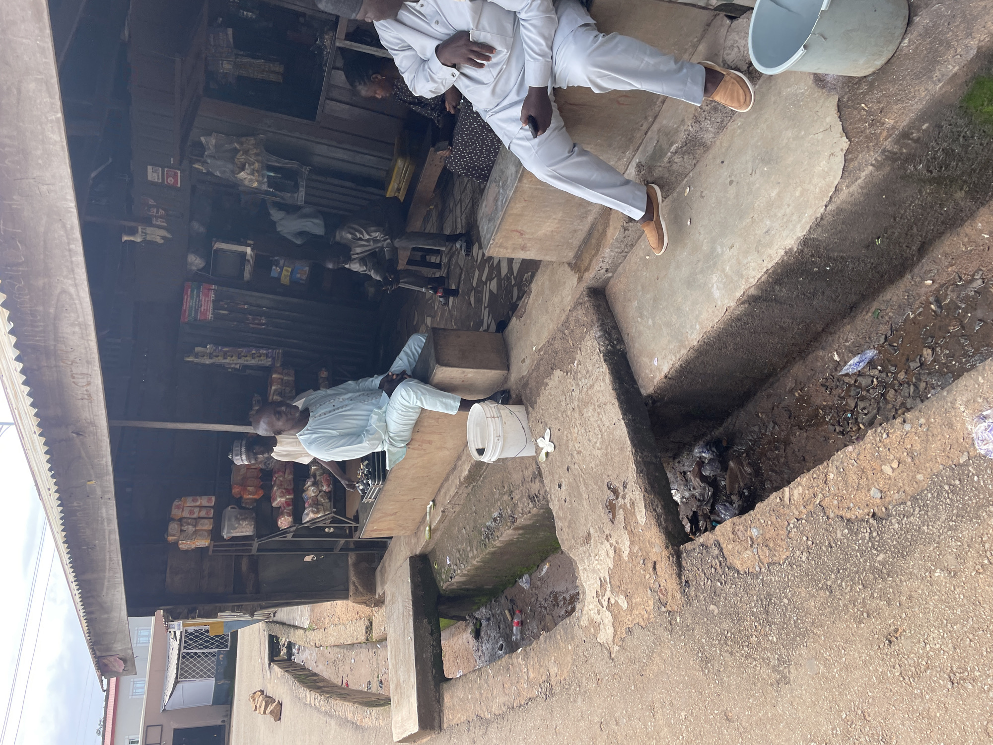 The image depicts an outdoor scene in Jos, Nigeria, featuring a makeshift market stall. In the foreground, a concrete drainage channel runs alongside a dirt path. A white bucket and a flower are visible near the channel.
Several individuals are present. Two men in traditional light blue attire are seated on a raised concrete platform in front of the stall. One is holding a mobile phone. Another man, wearing a dark cap and traditional attire, is partially visible behind them, possibly interacting with goods on a shelf. Further back, a woman in a patterned dress is seated, and another man is visible in the background.
The stall itself is constructed from wood and corrugated metal. Shelves are stocked with various packaged goods, including what appear to be bread loaves and smaller items. A small television is mounted on a wall inside the stall. The overall environment appears to be utilitarian and somewhat unkempt, with visible debris in the drainage.