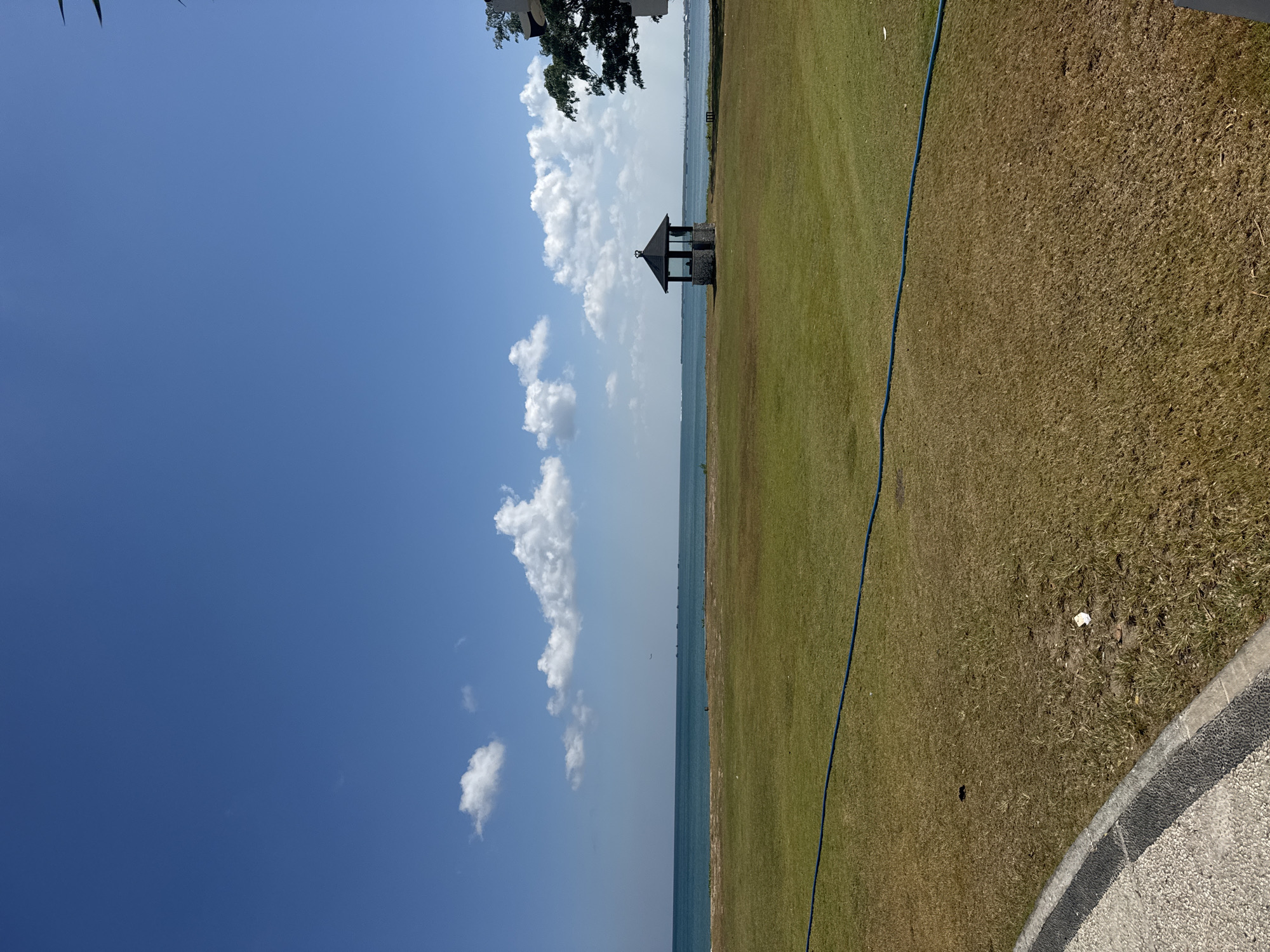 This image depicts a scene in Kuta Selatan, Indonesia, characterized by a vast expanse of green grass leading to a tranquil ocean under a clear blue sky dotted with white clouds. In the middle ground, a small, octagonal gazebo with a stone base and a dark, pointed roof stands near the water's edge. The ocean stretches to the horizon, where a faint line of land or distant structures can be seen. A blue hose lies horizontally across the lawn in the foreground. To the far right, the corner of a building and the lush green leaves of a tree are visible. The overall environment is serene and natural, suggesting a public park or resort area.