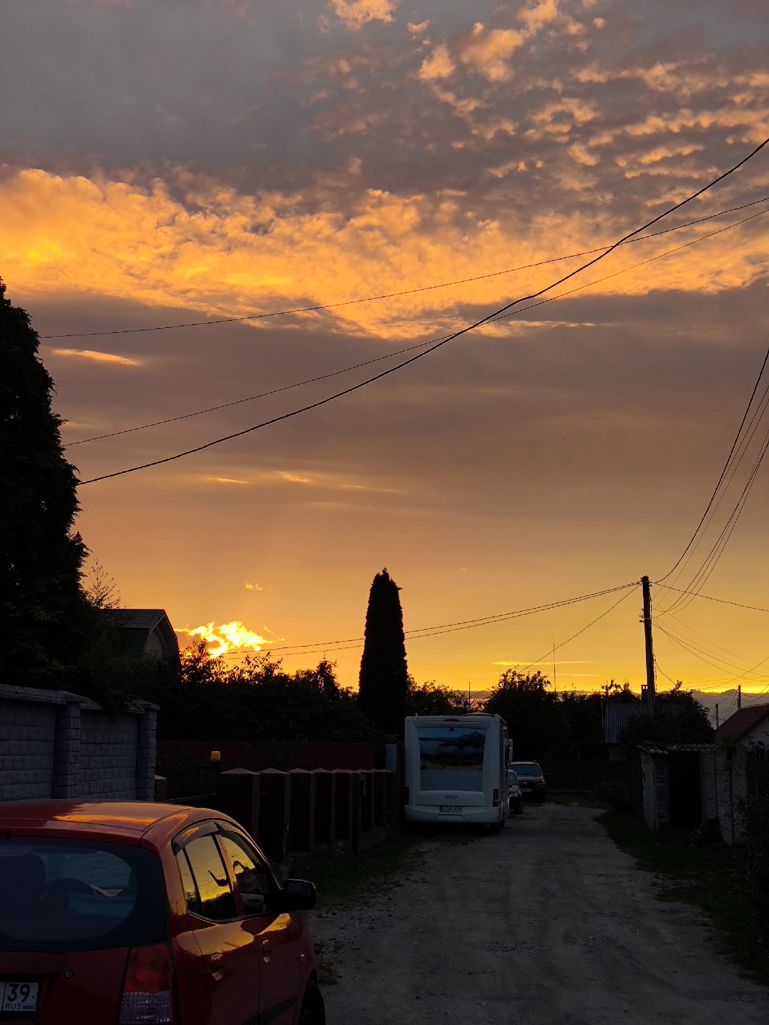 This image depicts a dirt road in a residential area during sunset. The sky is dominated by vibrant orange and yellow hues, with scattered clouds illuminated by the setting sun. In the foreground, the rear of a red car is visible on the left side of the road, reflecting the sunset on its surface. Further down the road, a white RV is parked. A tall, dark, conical tree stands to the right of the RV, and a utility pole with multiple wires is also visible on the right side of the road. Various houses and fences are present throughout the scene, suggesting a suburban or rural setting. The overall impression is of a peaceful evening in a quiet neighborhood. The license plate on the red car indicates it is registered in Kaliningrad Oblast, Russia.