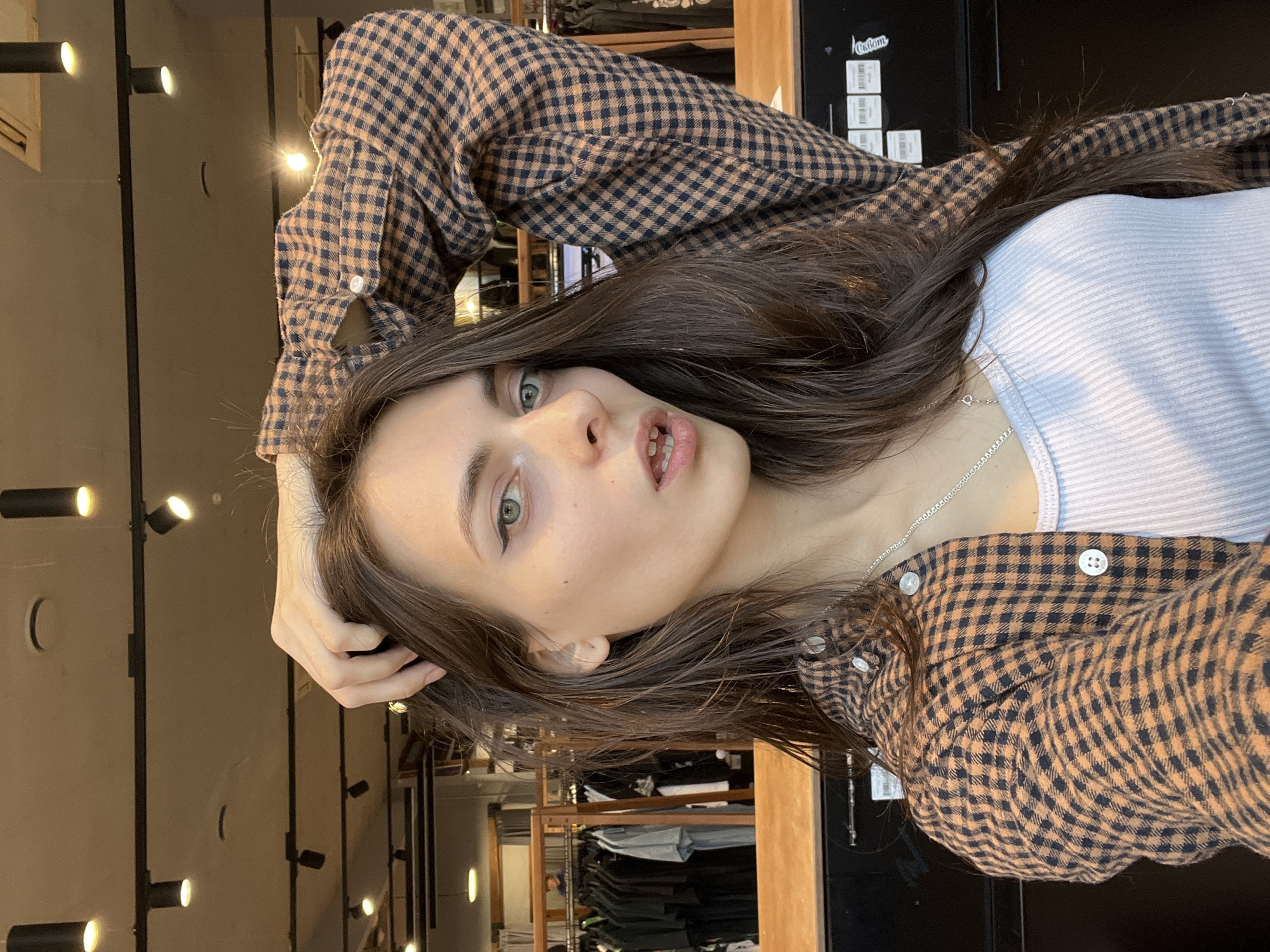 A young woman poses indoors, presumably in a retail setting or store. She is wearing a white tank top layered under a brown and black checkered flannel shirt. Her dark hair is down, partially obscuring the tank top. She is wearing a silver necklace. Her left hand is raised to her head.
The background reveals clothing racks and fixtures typical of a store, suggesting the location might be within a shopping center or boutique. The lighting is artificial, featuring track lighting fixtures attached to the ceiling, common in commercial spaces. Based on the presence of a "Сибирские Зори" clothing store, the image may have been taken in Krasnoyarsk, Russia.
