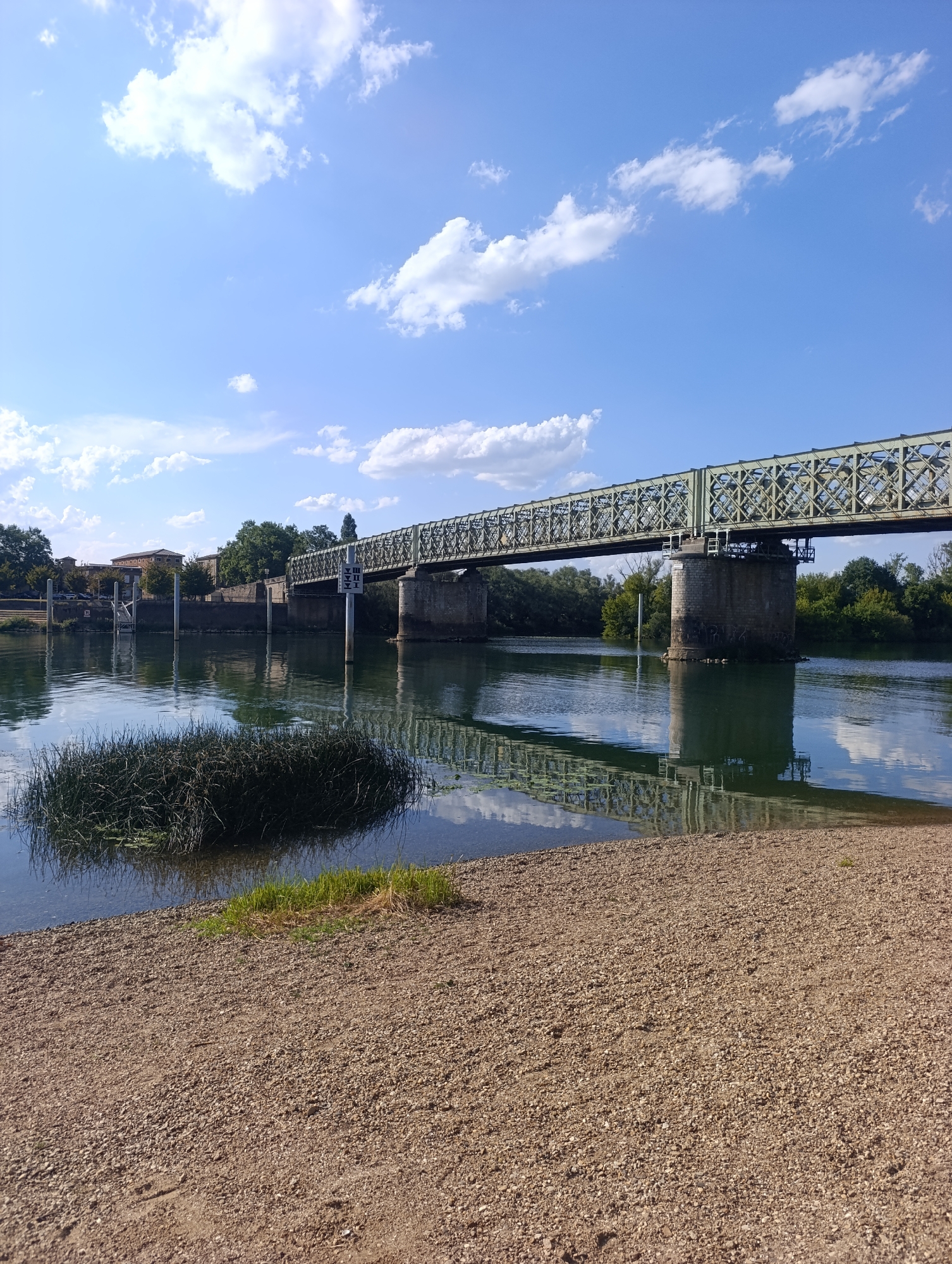 An eye-level, long shot captures a metal truss bridge spanning a calm river under a bright blue sky with scattered white clouds. The bridge, painted a muted green, is supported by stone pillars and shows a decorative lattice design. The river's surface reflects the bridge and the sky, with a small island of reeds in the foreground. The riverbank is covered in small stones and gravel. To the left of the bridge, buildings and trees are visible on the far bank. A navigational marker with directional arrows stands in the river near the bridge. The scene is set in Guéreins, France, near the Saône river.