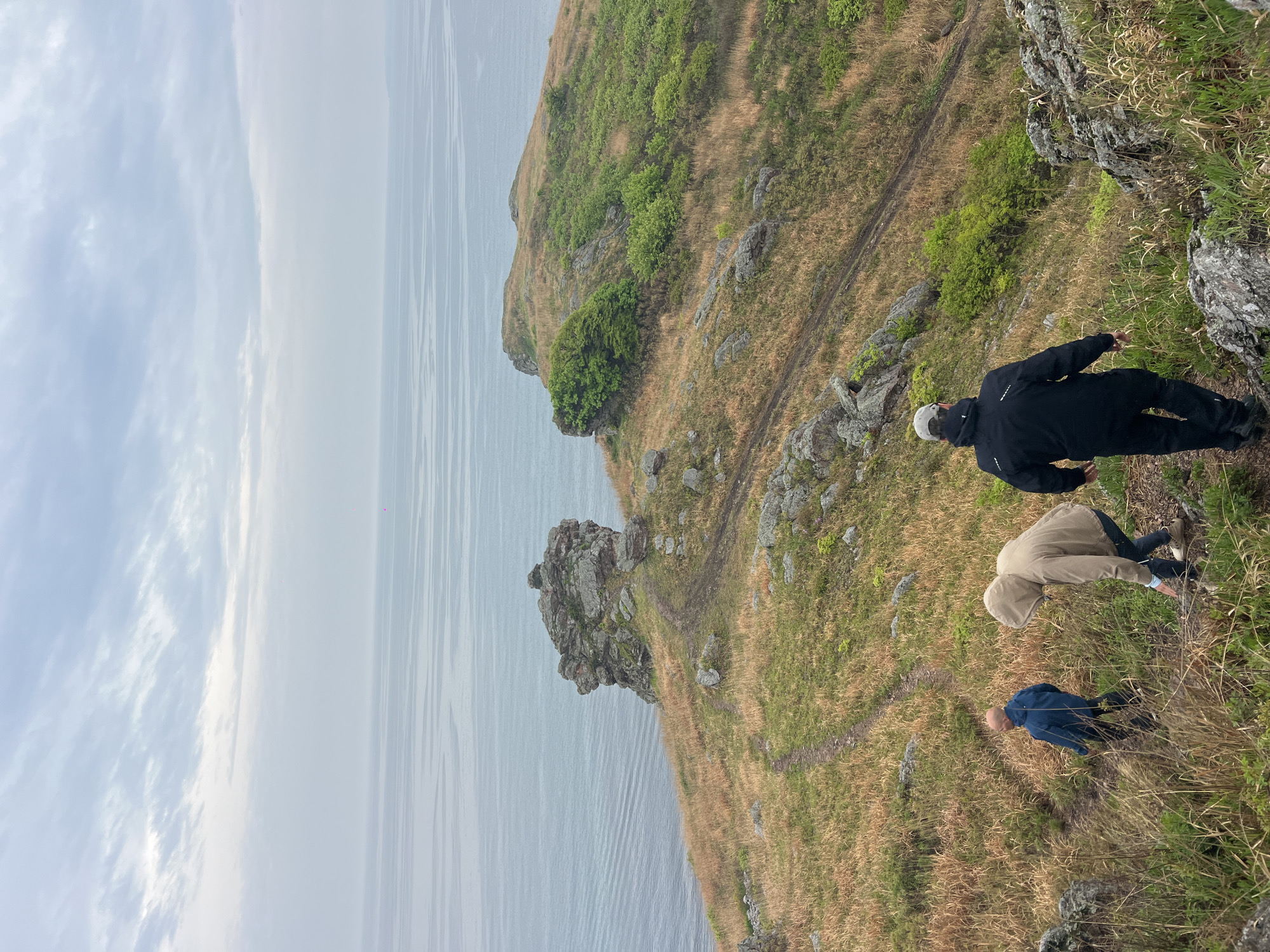 The image shows three people on a grassy cliff overlooking the ocean. A person in a black jacket and light cap is near the edge of the cliff on the right. Below them, another person is wearing a tan jacket with a hood. Further down the slope is a person in a blue jacket. The landscape consists of dry grass, rocks, and patches of green vegetation. The sea is visible in the background, under an overcast sky. The location is Gvozdevo, Russia.