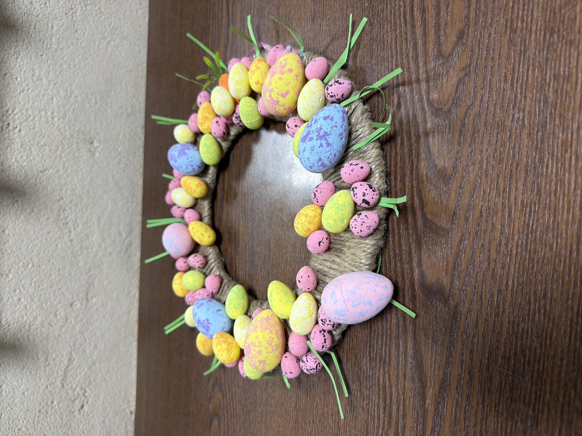 An Easter-themed wreath, adorned with numerous colorful, speckled eggs and green accents, hangs on a dark brown wooden door. The wreath's base is wrapped in natural jute rope or twine, providing a rustic texture.
The eggs vary in size and color, creating a vibrant display. Larger, elongated eggs are present in shades of pastel yellow, pink, and light blue, each speckled with contrasting colors (e.g., yellow with pink speckles, blue with purple speckles, pink with light blue speckles). Interspersed among these are smaller, more rounded eggs, primarily in pink with black speckles, and various shades of yellow, green, and orange. The eggs appear to be made of a lightweight material, possibly foam or plastic.
Small, thin strips of bright green plastic or artificial grass are strategically placed around and within the clusters of eggs, mimicking new spring growth.
The background consists of the dark brown wooden door, featuring a prominent vertical grain pattern, and to its left, a light-colored (off-white or beige), textured wall. The wreath is positioned slightly off-center, allowing both the door and a portion of the wall to be visible. The lighting is bright, highlighting the vivid colors and details of the festive decoration. No text is visible in the image.