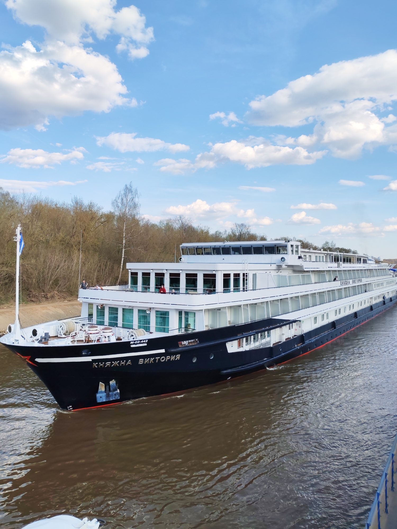 A full shot captures the "Knyazhna Viktoriya" river cruise ship in Varvarino, Russia. The ship is a multi-decked vessel with a black hull and white upper levels. The name of the ship, "КНЯЖНА ВИКТОРИЯ," is displayed on the front of the hull in gold lettering. A flag is hoisted at the front.
The ship is sailing on a wide river with brown water. The riverbank is visible on the left side, covered with trees that have not yet fully leafed out. The sky above is blue with scattered white clouds. The foreground is a portion of another ship.