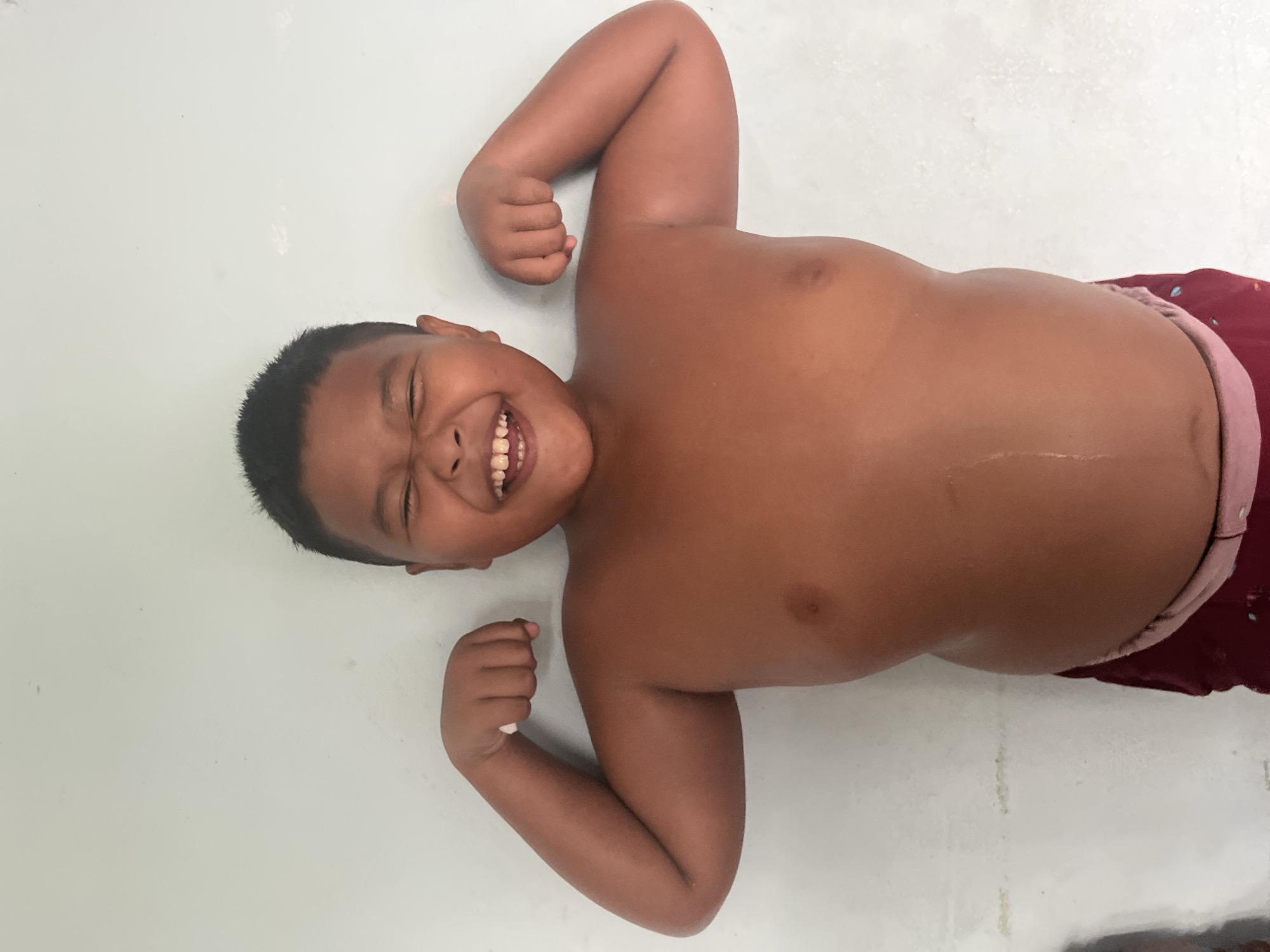 A young boy with dark hair and brown skin smiles with his eyes closed while flexing his biceps. He is wearing red shorts. The boy is standing in front of a light-colored wall.