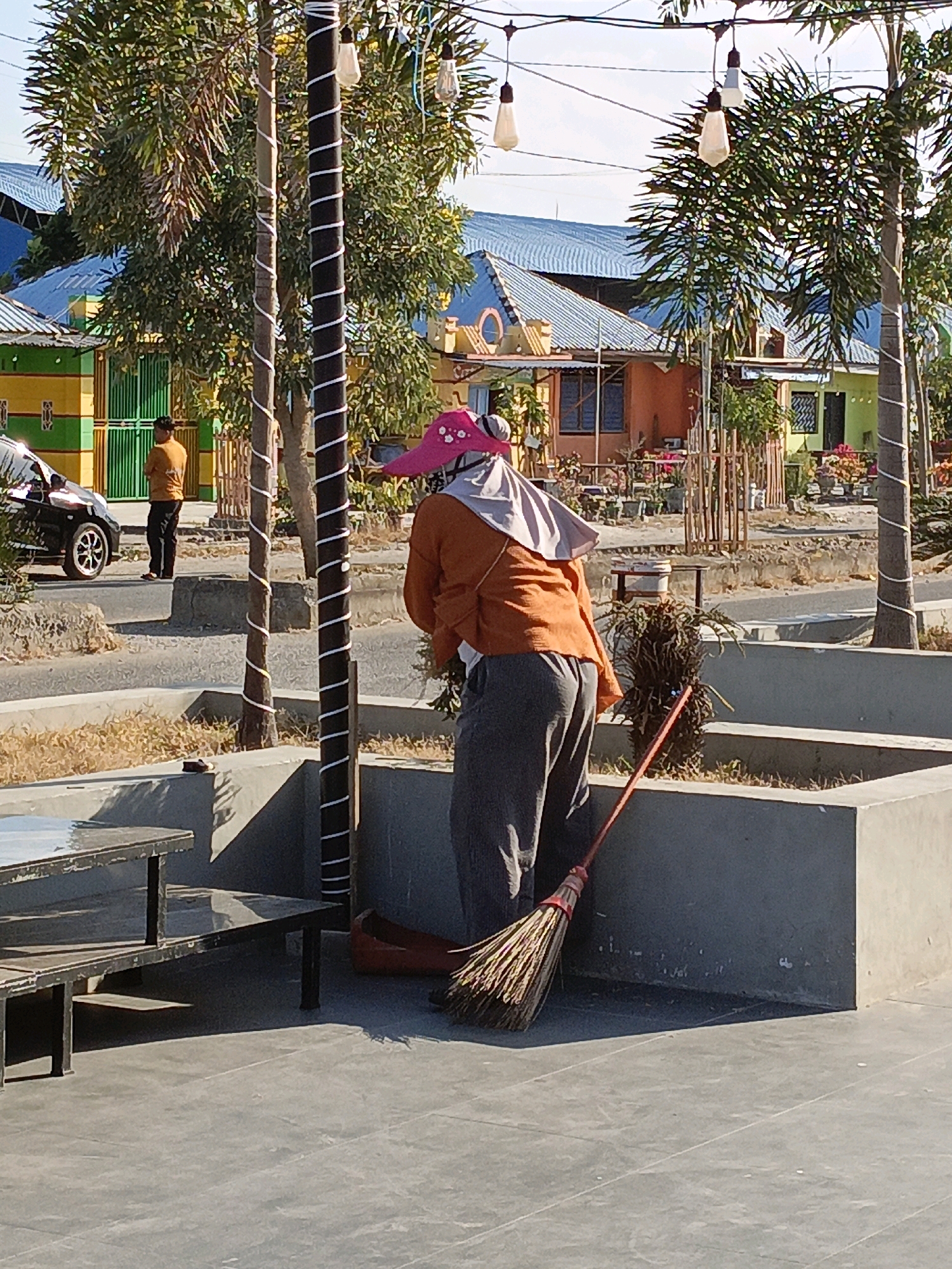 In Ela-ela, Indonesia, a person stands facing away from the camera, holding a broom in a concrete outdoor area. The individual is wearing an orange top, gray pants, and a pink wide-brimmed hat, with a light-colored cloth draped over their head. They are positioned next to a tall, dark pole wrapped with white tape, and a broom is leaning against a concrete planter box behind them, with its bristles touching the ground. In the background, colorful buildings with blue roofs are visible, along with palm trees and a black car parked on a road. A person in an orange shirt stands near the car, looking towards the buildings. The ground is paved with gray tiles in the foreground and gravel in the mid-ground. The scene is illuminated by daylight.