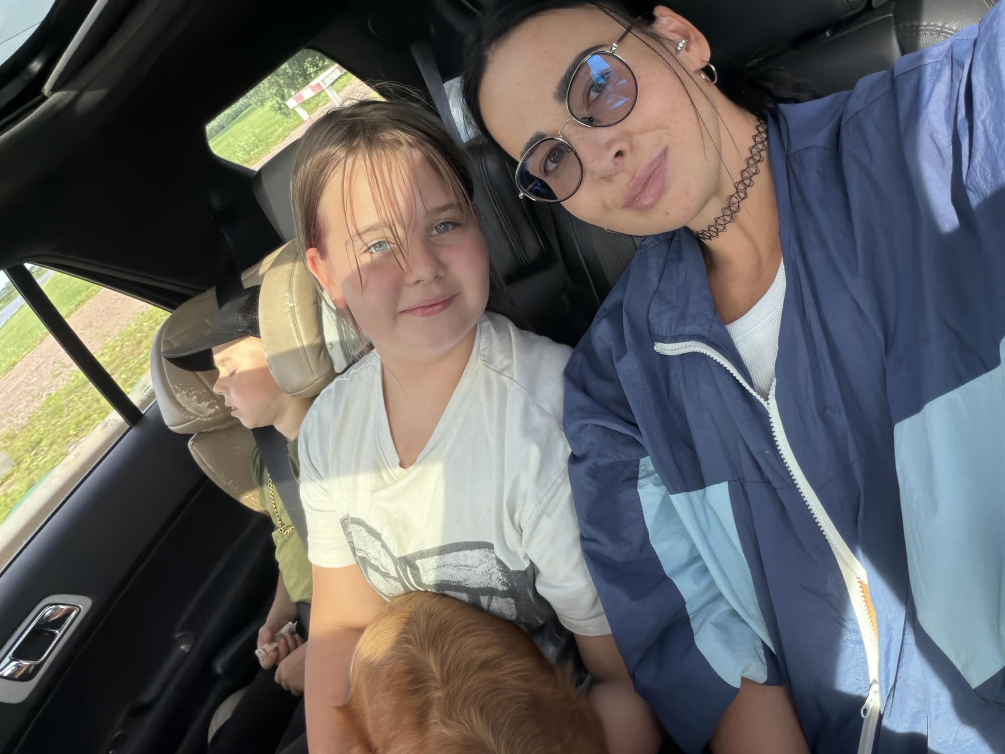 In the interior of a vehicle, three individuals, two children and one adult female, are seated. The adult female is in the foreground, looking towards the camera, and is wearing a blue and light blue jacket over a white top, with eyeglasses and a black choker necklace. To her left and slightly behind her, a young girl is looking towards the camera with a smile, wearing a white t-shirt. Further back and to the left, a younger child is visible, appearing to be asleep or resting, wearing a green shirt. A dog with tan fur is partially visible, nestled beside the young girl. The background shows a glimpse of a rural landscape with fields and vegetation through the car windows. The location is identified as the city of Шолохово, Russia.