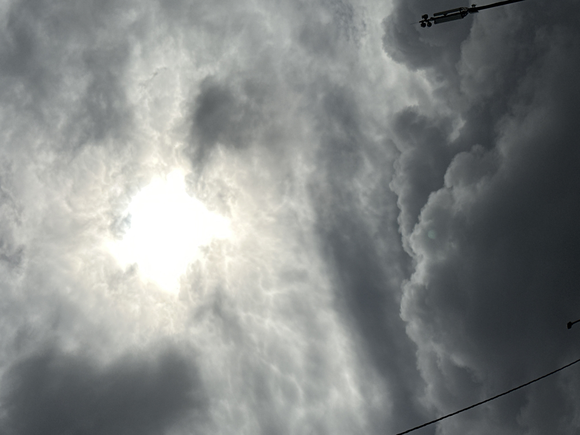 The image shows a dramatic sky with thick, dark clouds partially obscuring the sun. The sun is visible as a bright, almost white light source in the upper center of the frame, with its rays spreading outwards. The clouds vary in density and shade, with some areas appearing almost black and others a lighter gray, creating a textured and turbulent effect. In the lower right corner, a tall, slender pole with a wind sensor and antenna array is visible against the dark clouds. A dark, thin wire runs diagonally from the upper left towards the center of the image. The overall impression is one of an overcast or stormy sky. The location is the city of Podolsk, Russia.