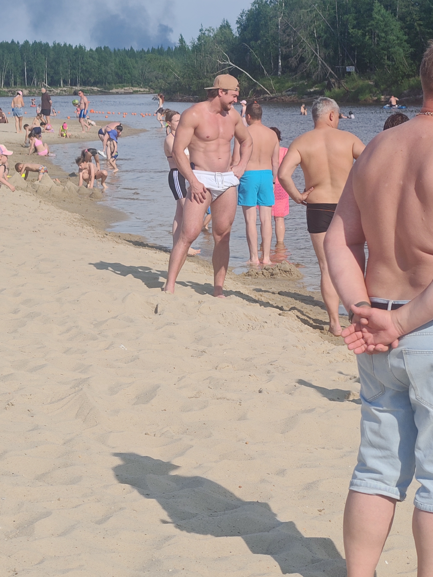 A group of people are on a sandy beach with a body of water in the background. The water is calm and reflects the sky, which is partly cloudy. Dense green forest lines the far bank of the water. Several people are in the water, and others are on the beach. Some individuals are sunbathing, while others are playing in the sand. A few people are standing in the shallow water. The overall scene suggests a warm, sunny day at a popular recreational area. The location is the beach of the Seyakha River in Novy Urengoy, Russia.