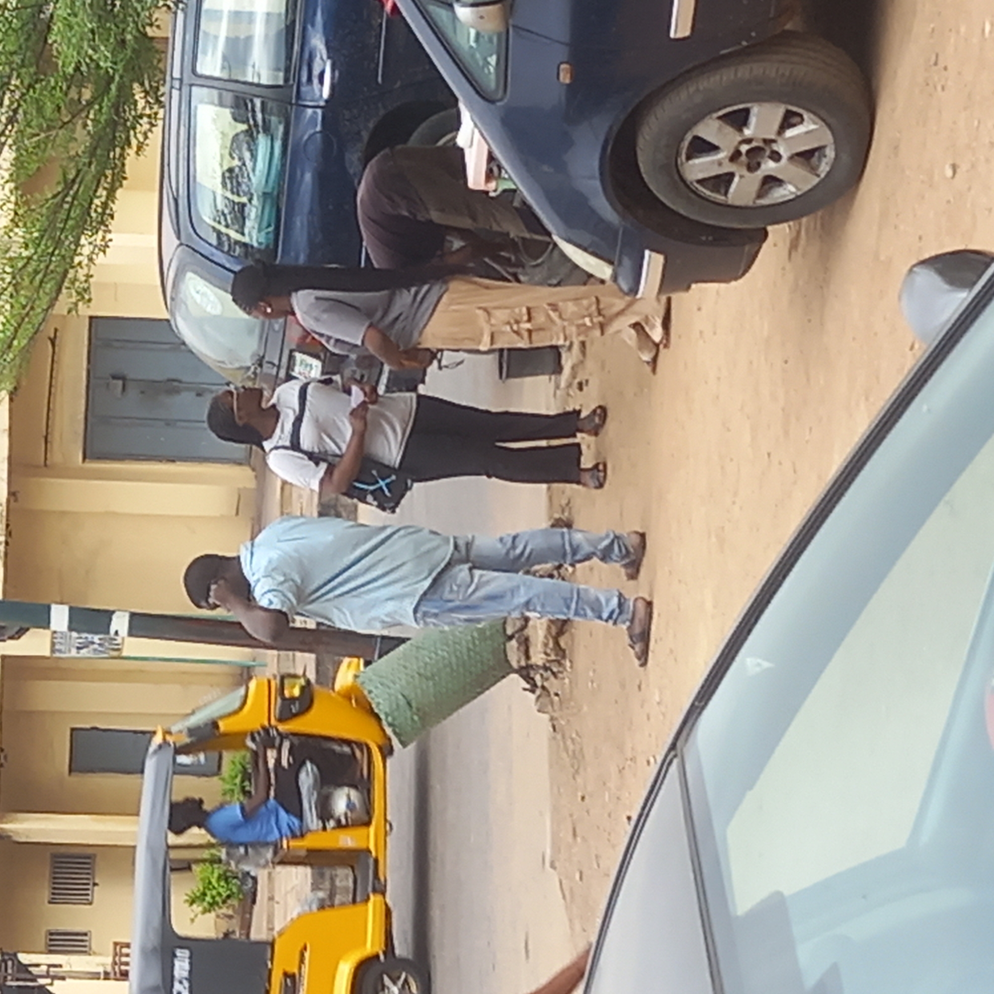 The image shows a street scene in Dadi Riba, Nigeria. A blue minivan is parked on the side of the road, and a man in dark clothing is working on its undercarriage. Two other people, one wearing a white shirt and black pants and another wearing a gray shirt and tan pants, stand nearby, possibly observing the work. Another man in a blue shirt and jeans stands slightly apart. A yellow motorized tricycle taxi is parked at the curb, with a driver visible inside. Parts of two cars are visible in the foreground on the side of the camera. The background features a light yellow building with closed doors and windows. The surface looks to be dirt.