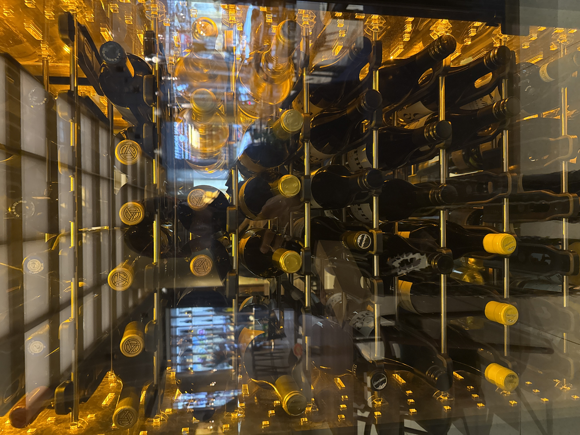 The image displays a head-on view of a glass-fronted wine cooler or display case, packed with numerous wine bottles arranged horizontally on multiple metal racks. The interior of the cooler is brightly lit with a warm, golden illumination. The bottles consist primarily of dark-colored glass, with some lighter, possibly transparent, glass bottles interspersed throughout. Visible bottle capsules are gold, yellow, and black. Several gold capsules feature a distinct circular emblem, and identifiable brands on labels or capsules include "MASSOLINO," "CANTINE LUNAE," "SUDKOL," and "CANTINE DEI MARRUCA." The reflective glass surface of the cooler shows the blurred interior of a restaurant or bar, including reflections of additional bottle displays, bar stools, and ambient lighting. A clear reflection of text on the glass identifies the establishment as "BERNACHE WINE & TAPAS." This location is Bernache Wine & Tapas, situated at ul. "Paris" 1A, 1000 Sofia Center, Sofia, Bulgaria. No individuals or active interactions are depicted in the scene.