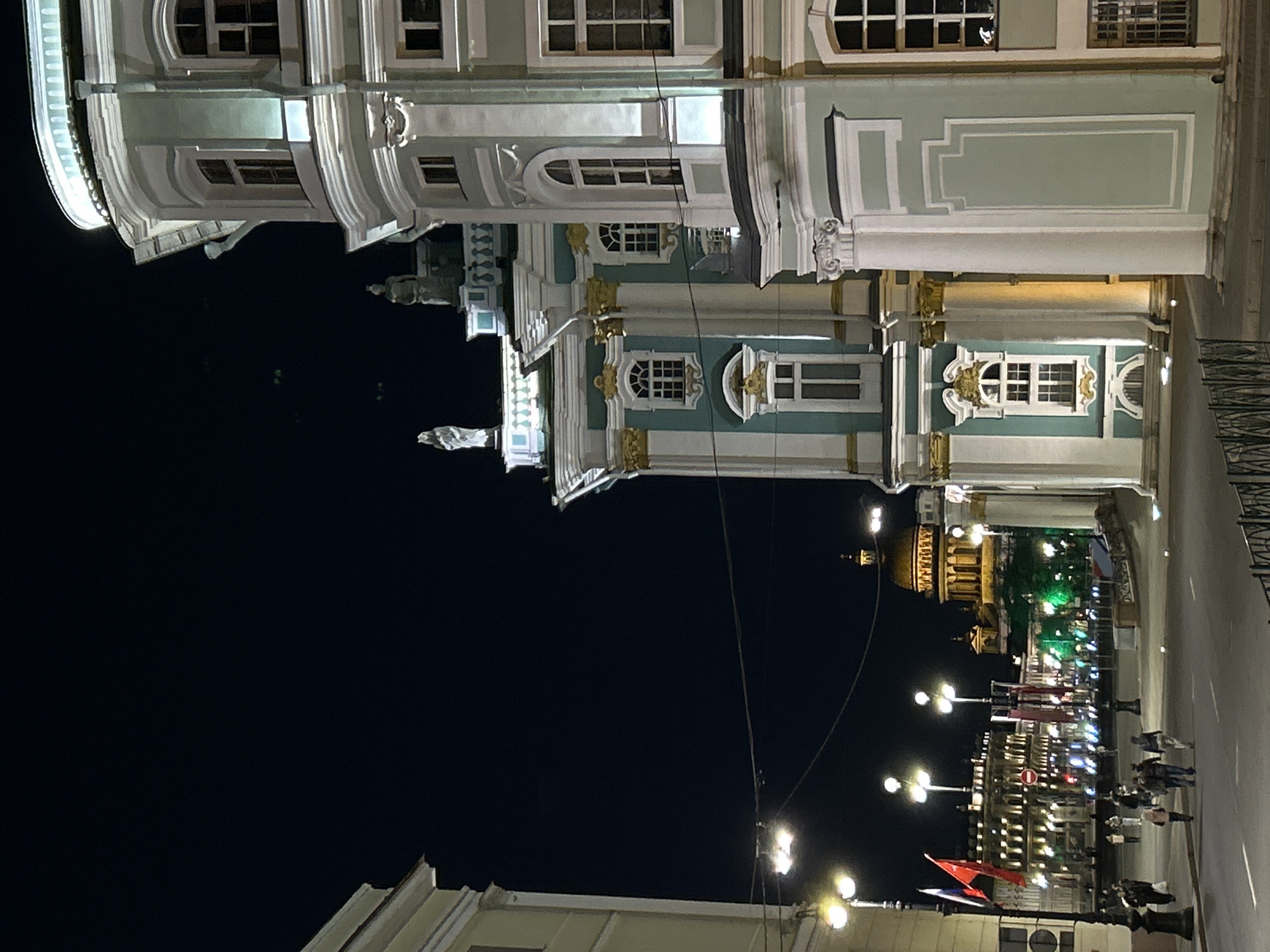 The image shows a nighttime scene in Saint Petersburg, Russia. The Winter Palace, part of the Hermitage Museum complex, is visible along the top half of the image. The building is illuminated and its distinctive architectural features are apparent. Below, a street scene unfolds with illuminated buildings and streetlights. Pedestrians are walking along the pavement. The area is lit by streetlights. In the middle-distance is St. Isaac's Cathedral.