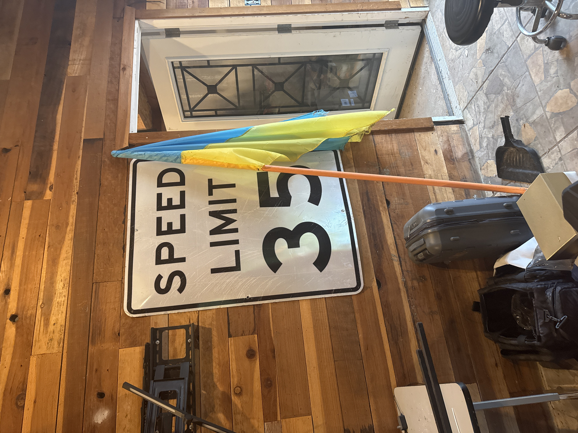 The image depicts an indoor scene with wooden paneled walls. A speed limit sign displaying "SPEED LIMIT 35" is prominently mounted on the wall. To the right of the sign, a Ukrainian flag on an orange pole is visible, partially obscuring a white door with a decorative glass insert. On the floor, several items are present, including a dark suitcase, a black backpack, a shovel, a stool with wheels, and a low table. The floor appears to be tiled. The scene suggests a storage or utility area, possibly a garage or workshop. The image is taken from a slightly low angle, looking upwards at the sign and flag. The lighting is moderate, with natural light possibly entering from the doorway. The presence of the Ukrainian flag suggests a connection to Ukraine or support for the country. The overall environment appears to be somewhat cluttered but organized. The location is identified as being in the city of Orangevale, United States.