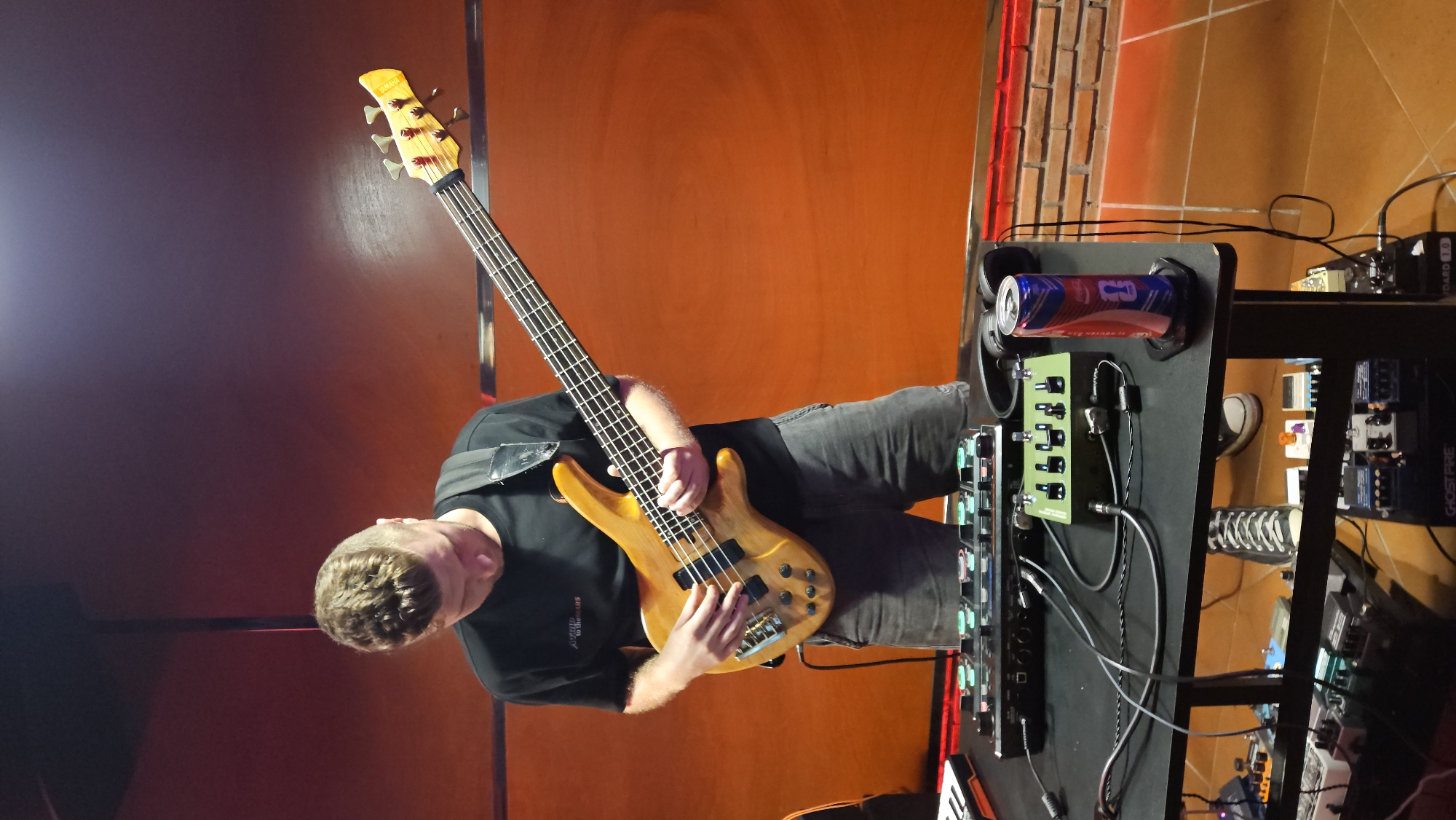 A person is playing a yellow bass guitar, standing next to a black equipment rack with musical effects pedals, a can of Monster energy drink, and headphones. The location appears to be a performance venue with an orange wall and some brickwork. The location might be in Bắc Nha Trang, Vietnam.