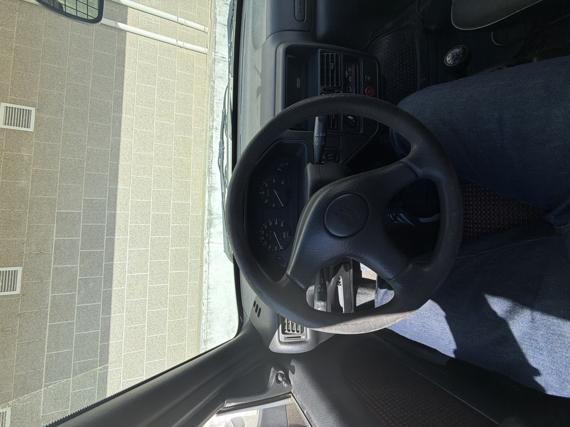 The image depicts the interior of a car, specifically the dashboard and steering wheel, as viewed from the driver's seat. The steering wheel has a Peugeot logo in the center. The driver's legs, clothed in dark blue jeans, are visible. Outside the car window, there is a paved area with visible storm drains and adjacent building. Based on the interior details, the vehicle is likely an older Peugeot. The setting appears to be an urban or suburban environment. There is no explicit information within the image that places the location in Vila Real, Portugal; this needs to be identified by external contextual information. There are no other people visible, and the action depicted is unclear, however, the image is potentially depicting the inside of a car while it is parked.
