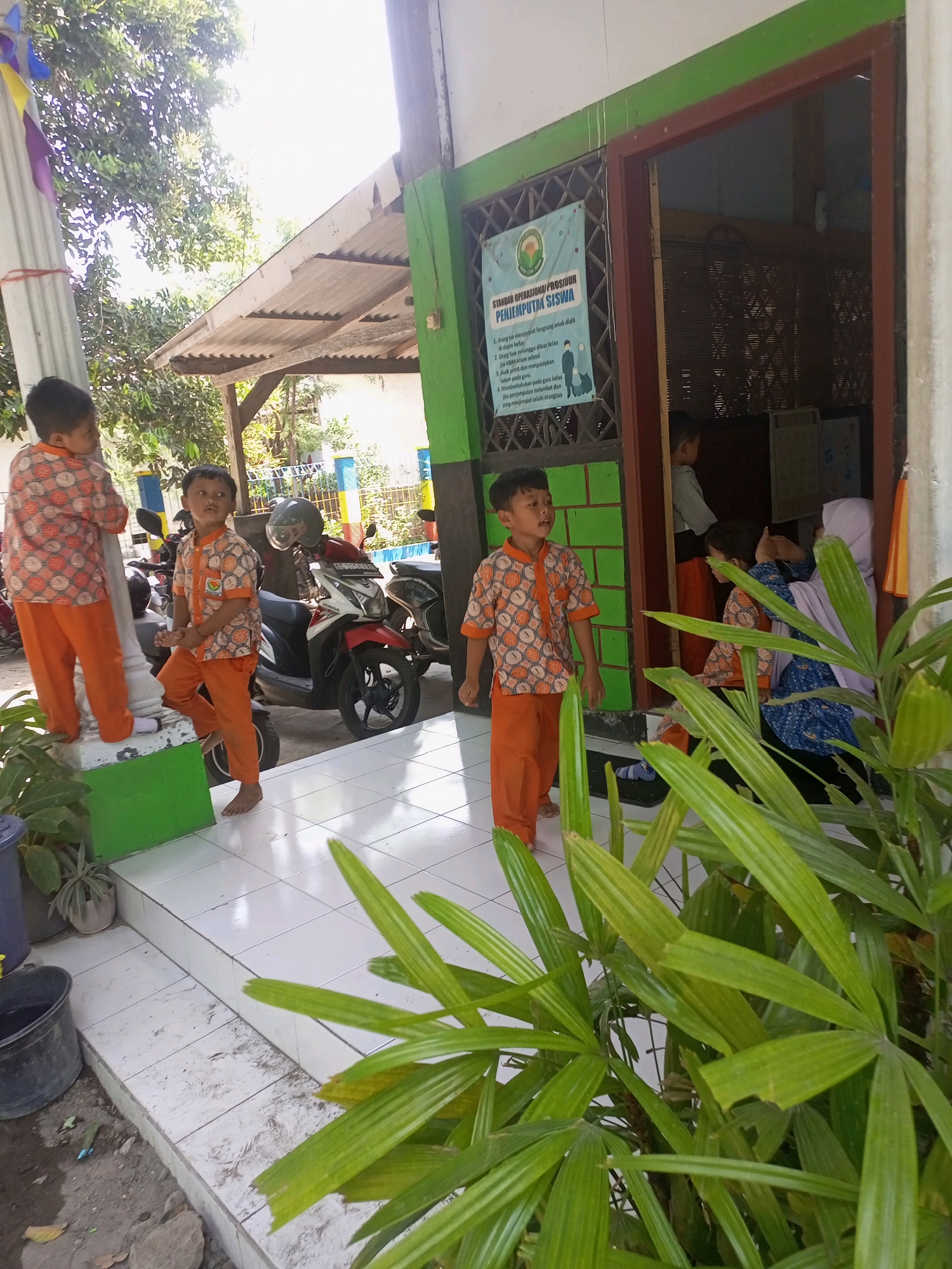 In the city of Kediri, Indonesia, a group of children, presumably students, are gathered outside a building. Three boys in matching orange pants and patterned shirts are the most visible figures. One boy stands on a raised platform, another leans against a pillar, and a third stands to the right of the frame. Behind them, a few motorcycles are parked. The building's exterior features green and brown accents, with an open doorway revealing more figures inside, including a woman wearing a hijab. A poster with text in Indonesian is affixed to the green wall near the entrance. The scene appears to be outdoors, with trees and bright sunlight indicating daytime. A leafy plant is prominent in the foreground, partially obscuring the view. The ground is tiled, with some dirt and debris visible at the edge of the platform.