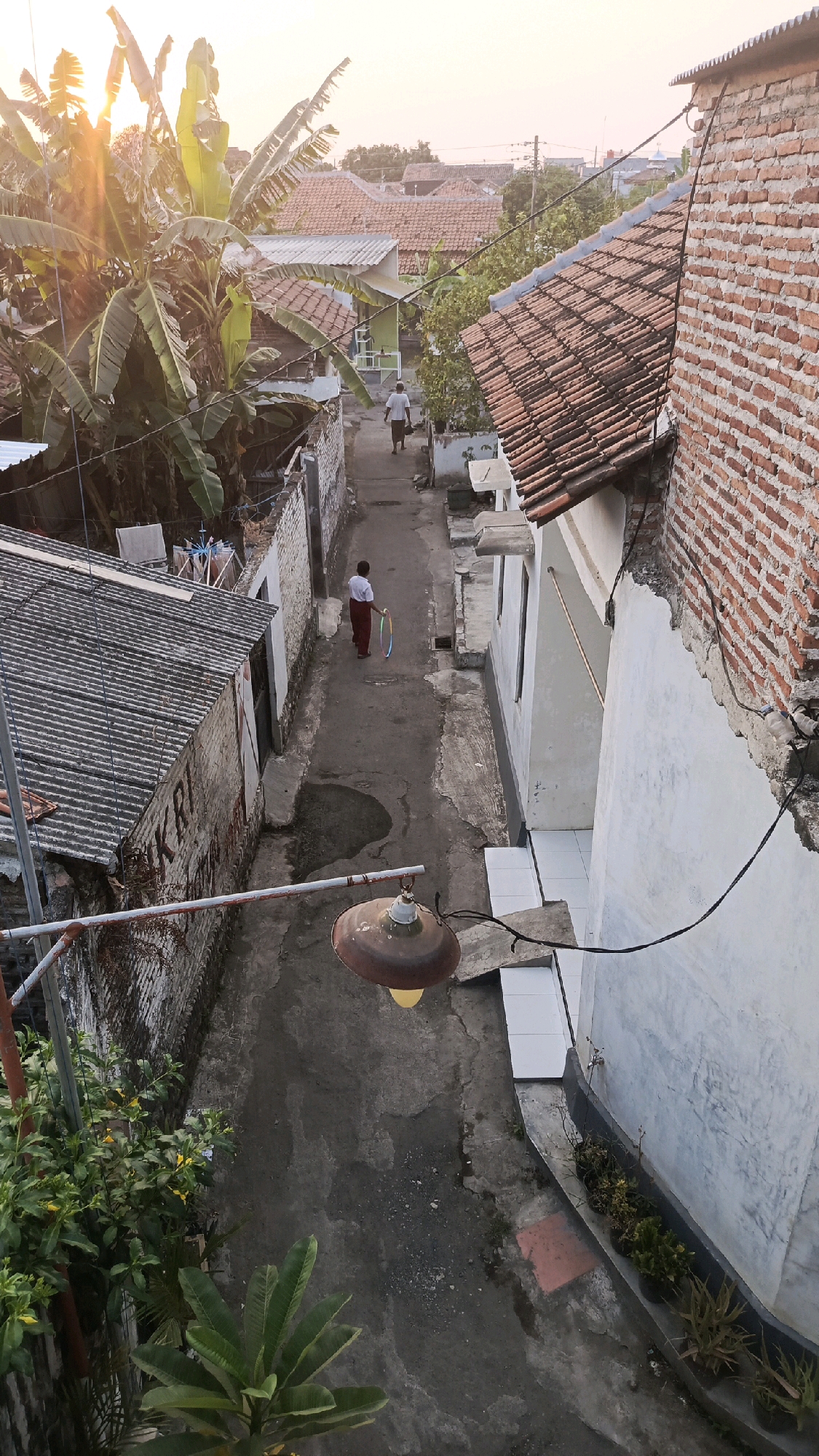 The image depicts a narrow alleyway between residential buildings in Pekauman Kulon, Indonesia. In the foreground, a rusted outdoor light fixture hangs from a metal pole. Lush green plants are visible on the left side of the frame. The alley itself is paved with concrete, showing signs of wear and tear.
Further down the alley, a young person, viewed from behind, is walking away from the camera, holding a colorful hula hoop. The person is wearing a white top and dark pants. Another individual, also viewed from behind, is walking further up the alley, away from the young person.
The buildings lining the alley are constructed with brick and concrete, some featuring exposed brickwork. Roofs are predominantly tiled with terracotta. Banana trees and other vegetation are interspersed among the houses, suggesting a tropical or subtropical environment. The setting sun casts a warm, golden light across the scene, illuminating the banana leaves and creating long shadows. The overall impression is of a lived-in, local neighborhood.