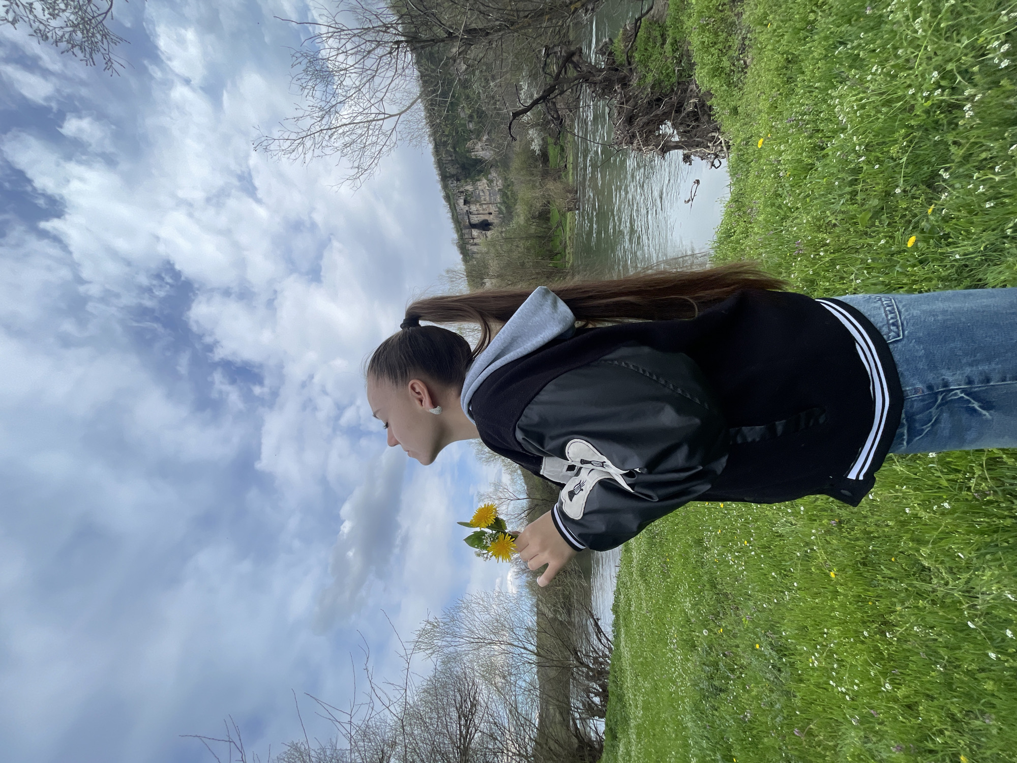 A young woman stands in a grassy area by a river. She has long brown hair pulled back in a ponytail and is wearing jeans and a black jacket with white stripes on the sleeves. She is holding a small bunch of yellow dandelions. The river flows in the background, with a cliff visible on the opposite bank. The sky is cloudy with patches of blue. The scene is the city of Barkach, Bulgaria.