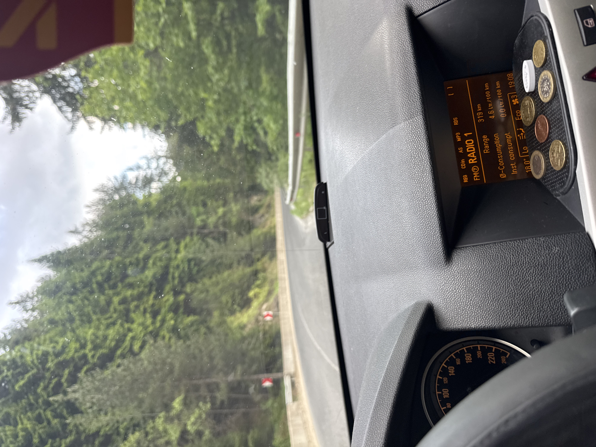 The image provides an interior view from a car, looking out the windshield at a road cutting through a dense, lush green forest. The setting appears to be a natural, possibly mountainous or hilly area, aligning with the location context of Sokolovtsi, Bulgaria. The sky is partially cloudy, indicating daytime with good ambient light, despite the time displayed.

Inside the vehicle, a digital display panel is prominently visible on the dashboard. It shows "FM RADIO 1", "Range 319 km", "Ø-Consumption 4.6 Ltr./100 km", and "Inst. consumpt. 0.0 Ltr./100 km". Other information includes "18.0°L" (likely 18.0 degrees Celsius), an "Eco" indicator, and the time "19:08". This time, coupled with the bright exterior, suggests late afternoon or early evening. Adjacent to the display, a coin holder contains various Euro coins, including 1 Euro, 50 cents, 20 cents, 10 cents, 5 cents, 2 cents, and 1 cent pieces. A small, white, oval object, possibly a pill or mint, is also present among the coins. A portion of the speedometer, marked from 80 to 240, is visible on the lower right, partially obscured by the steering wheel.

Outside the car, the asphalt road curves through the dense forest. Red and white chevron signs, commonly used in Europe to warn drivers of sharp bends, are visible further down the road, indicating a winding route. The activity captured is likely driving or having just stopped along a scenic forested road.
