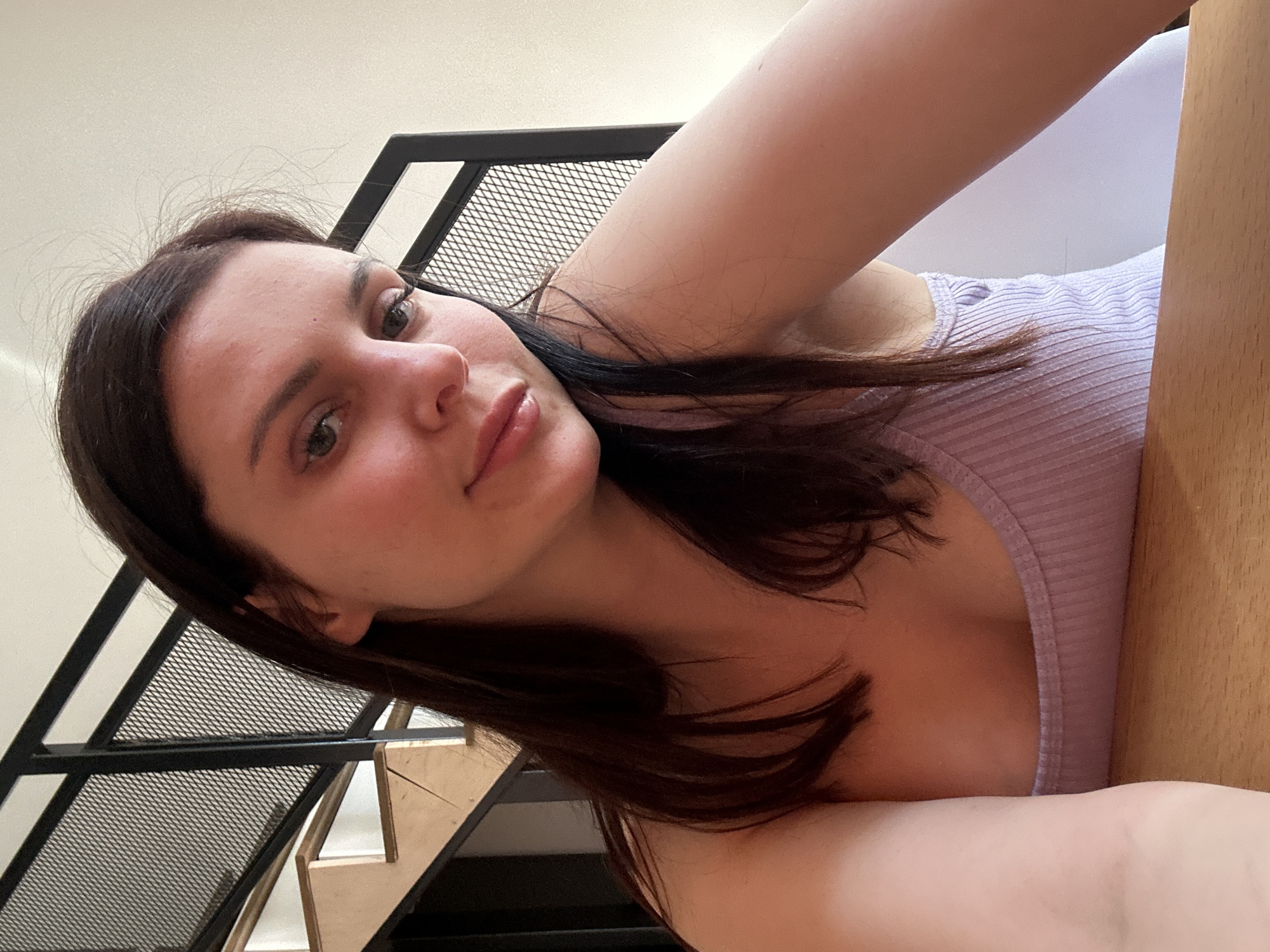 A young woman with dark brown hair and fair skin is shown in a close-up selfie. She is wearing a lavender ribbed tank top and is seated at a wooden table. Behind her, a modern staircase with black metal railings and wooden steps is visible. The background appears to be an indoor setting with white walls. Her expression is relaxed, with her head tilted slightly.