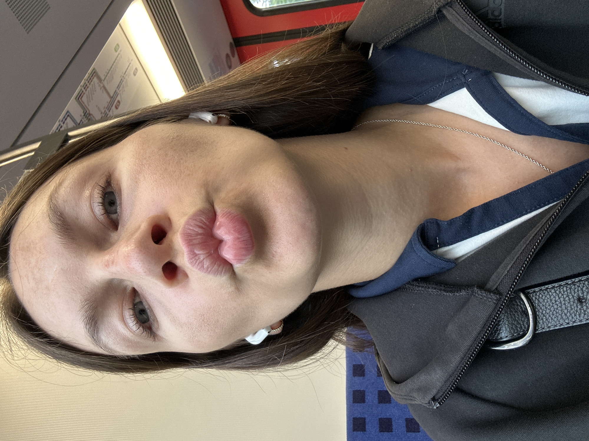 A close-up selfie captures a young woman inside what appears to be a public transport vehicle, likely a train, with the image rotated 90 degrees. She has long, dark brown hair and is making a "kissing face" or "duck face" with puckered lips, while her blue-grey eyes are open and looking towards the viewer. Her complexion is fair, with some minor skin texture visible.
She is dressed in a dark, possibly black or navy blue, hooded jacket or sweatshirt with a zipper, which is open to reveal a navy blue collar with a white stripe underneath, and a white garment beneath that. A thin silver chain necklace is visible around her neck. White wireless earbuds are worn in both ears. On her side, a black belt with a silver buckle is present, possibly from a bag strap or an accessory worn over her clothing. A partial "Adidas" logo is visible on the shoulder of her jacket.
The background elements confirm the setting. In the upper part of the image (when oriented correctly), a light-colored ceiling with an overhead light fixture and a public transport route map are visible. A red-framed window shows a hint of green vegetation outside, suggesting it is daytime. The lower part of the image features blue upholstery with a square pattern, characteristic of train or bus seating. The overall scene is a casual, playful moment captured during travel, potentially in Buxtehude, Germany.