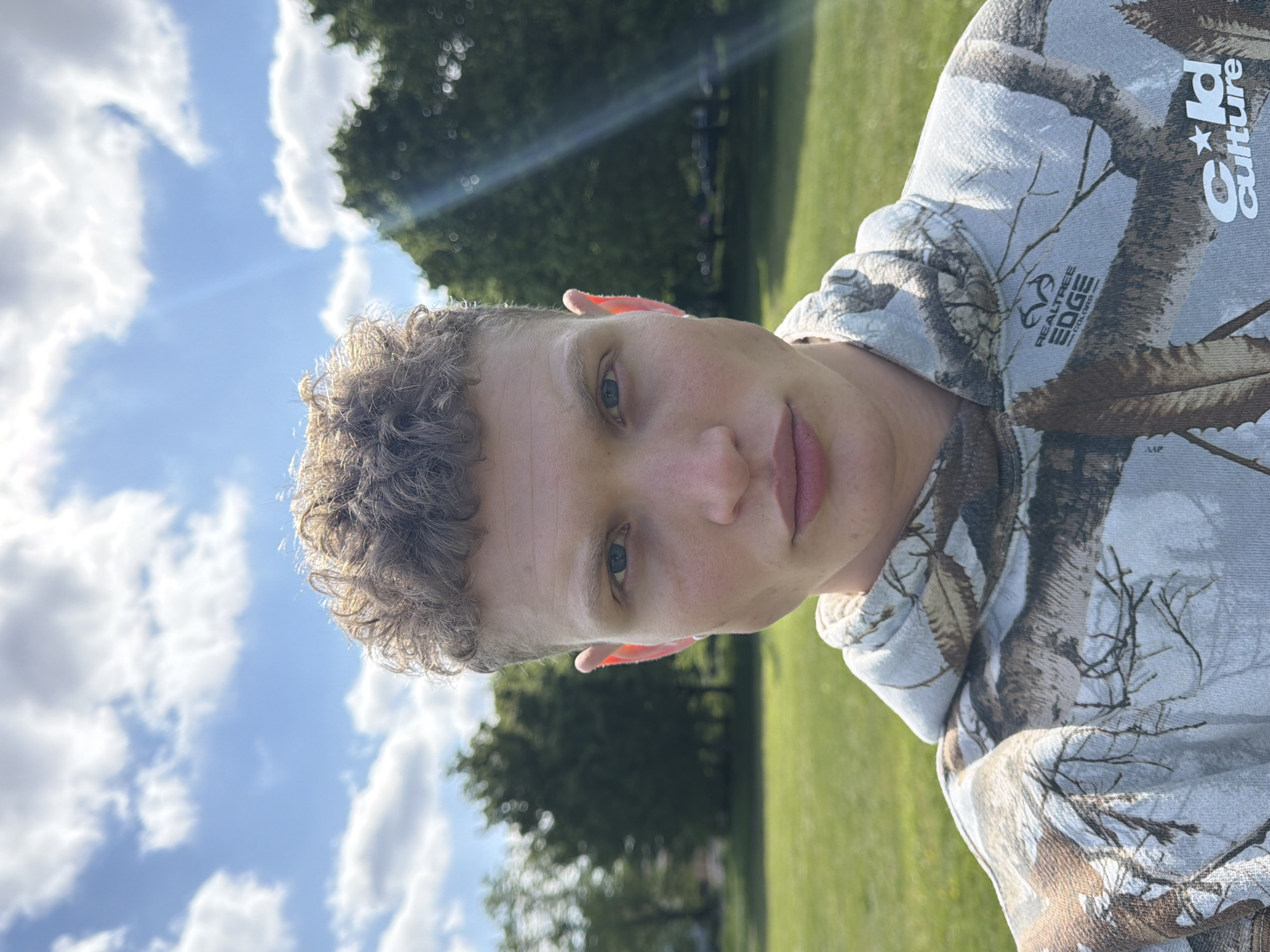 A close-up, upright portrait captures a young person with light skin, short, curly brown hair, and blue eyes, looking directly at the camera with a neutral expression. They are wearing a hooded sweatshirt featuring a camouflage pattern of light grey, white, and brown with leafless tree branches. Visible text on the sweatshirt includes "REALTREE EDGE" and "Gold Culture".
The setting is outdoors, likely a park or open green space in London, United Kingdom. The background shows a bright blue sky with scattered white, fluffy clouds. Green grass covers the lower and right parts of the background, and dark green trees are visible, some out of focus, suggesting distance. Bright sunlight is evident, casting highlights and creating visible sun rays in the upper left of the sky, indicating it is daytime, possibly morning or afternoon. The overall atmosphere appears sunny and clear.