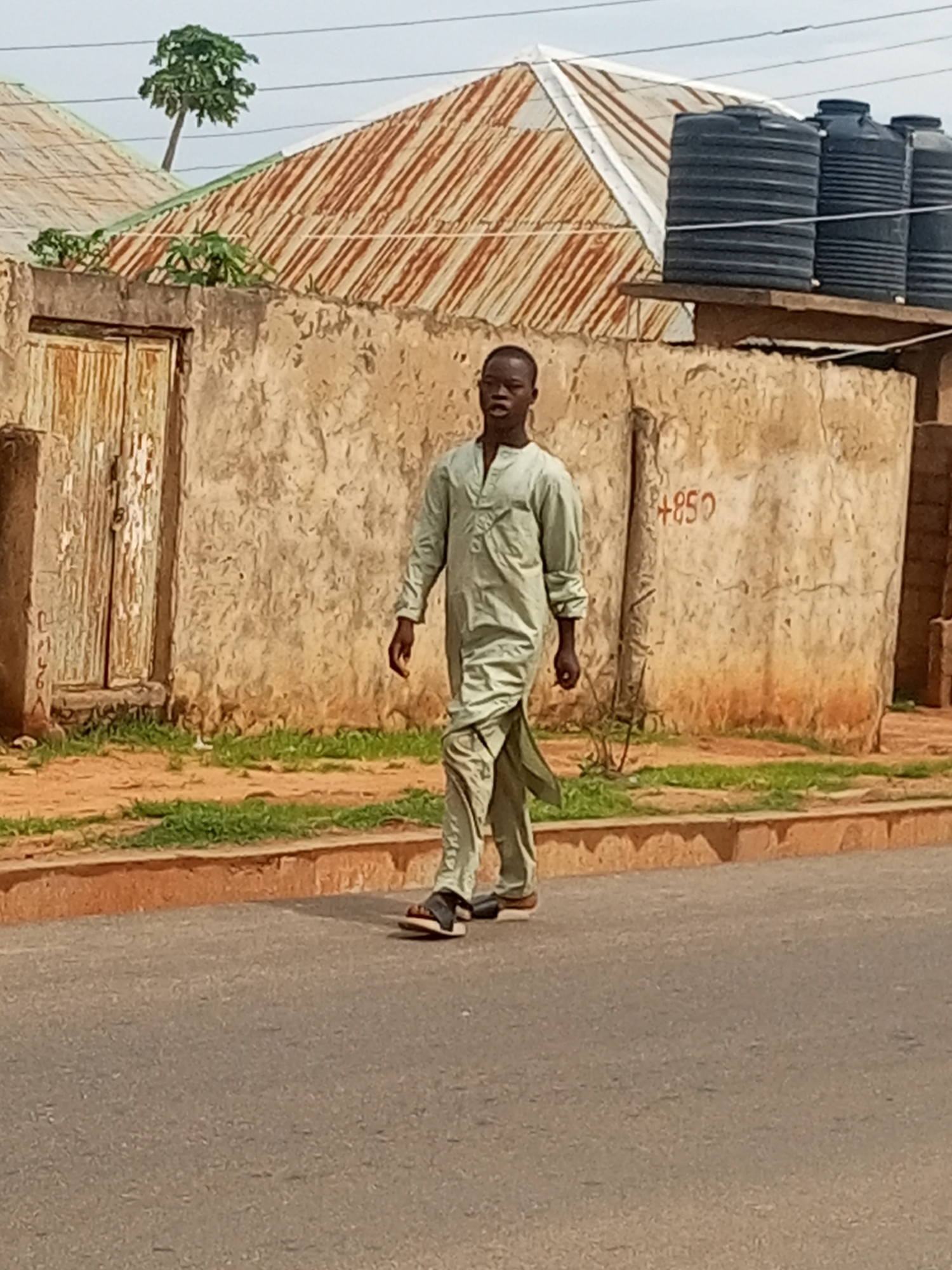 In Tempure, Nigeria, a young man walks along a paved road. He is wearing a light green, loose-fitting traditional outfit that consists of a tunic and trousers. He has dark skin and short dark hair, and his expression is neutral as he walks. The man is on the right side of the road, with the pavement in the foreground.
Behind him, a rough, textured wall is visible, painted in earthy tones of beige and orange. Painted on the wall are the numbers "+850" in red. To the left of the wall, a wooden door with peeling paint stands slightly ajar. Beyond the wall, two buildings with corrugated metal roofs are visible. One roof is a rusty orange, while the other is lighter, possibly white or silver. A small tree with green foliage grows out of the ridge of the rusty roof. To the right of the wall, three large black water tanks are stacked on a platform. The ground between the wall and the buildings is covered in sparse green grass and dirt. The sky is overcast.