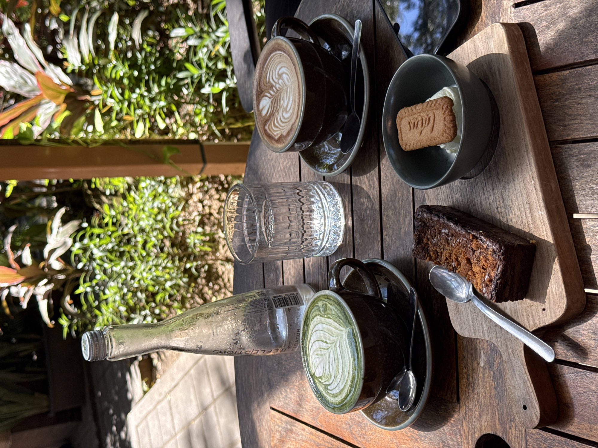 This outdoor scene depicts a wooden table in Kuta Selatan, Indonesia, arranged with a collection of beverages and dessert items. Two cups of coffee with latte art, one brown and one green, are presented on saucers with spoons. Adjacent to the coffees is a glass of water and a bottle laid horizontally. A bowl containing a biscuit atop a white substance, likely cream, sits alongside a piece of chocolate cake on a small wooden board. The scene is bathed in sunlight, with greenery visible in the background.