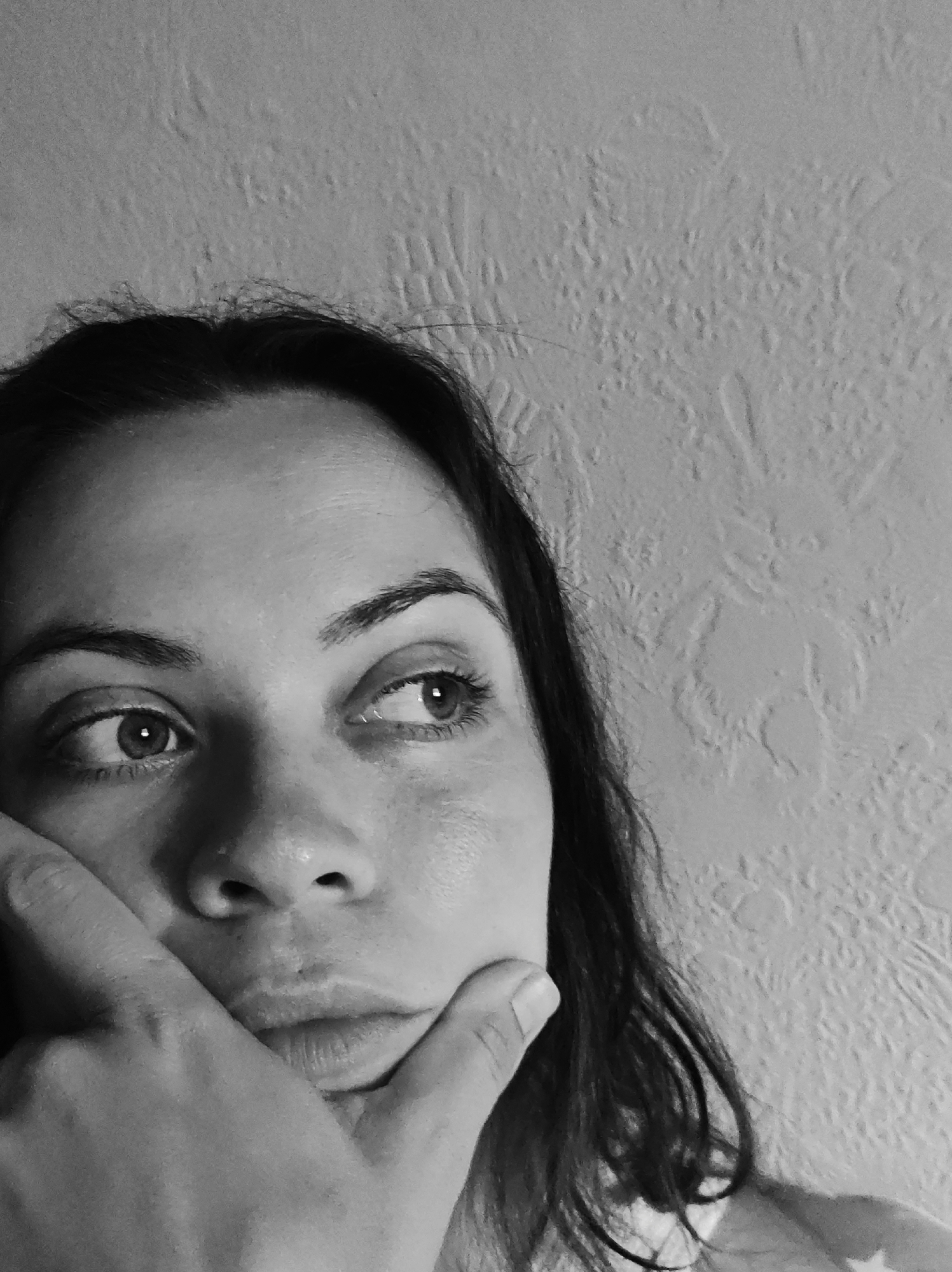 The image is a black and white close-up shot of a woman's face, cropped from the forehead down to her chin, with her left hand resting on her jaw. Her eyes are looking to her right, with a pensive expression. Her dark hair frames her face. The background is a textured wall, suggesting an indoor setting. The image does not contain any information about the city of Chaikovsky, Russia.